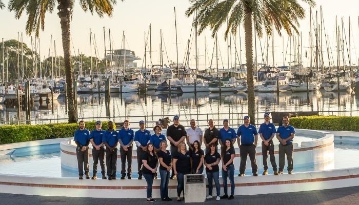 Group of people in blue and black shirts, posed in front of water fountain and boats.