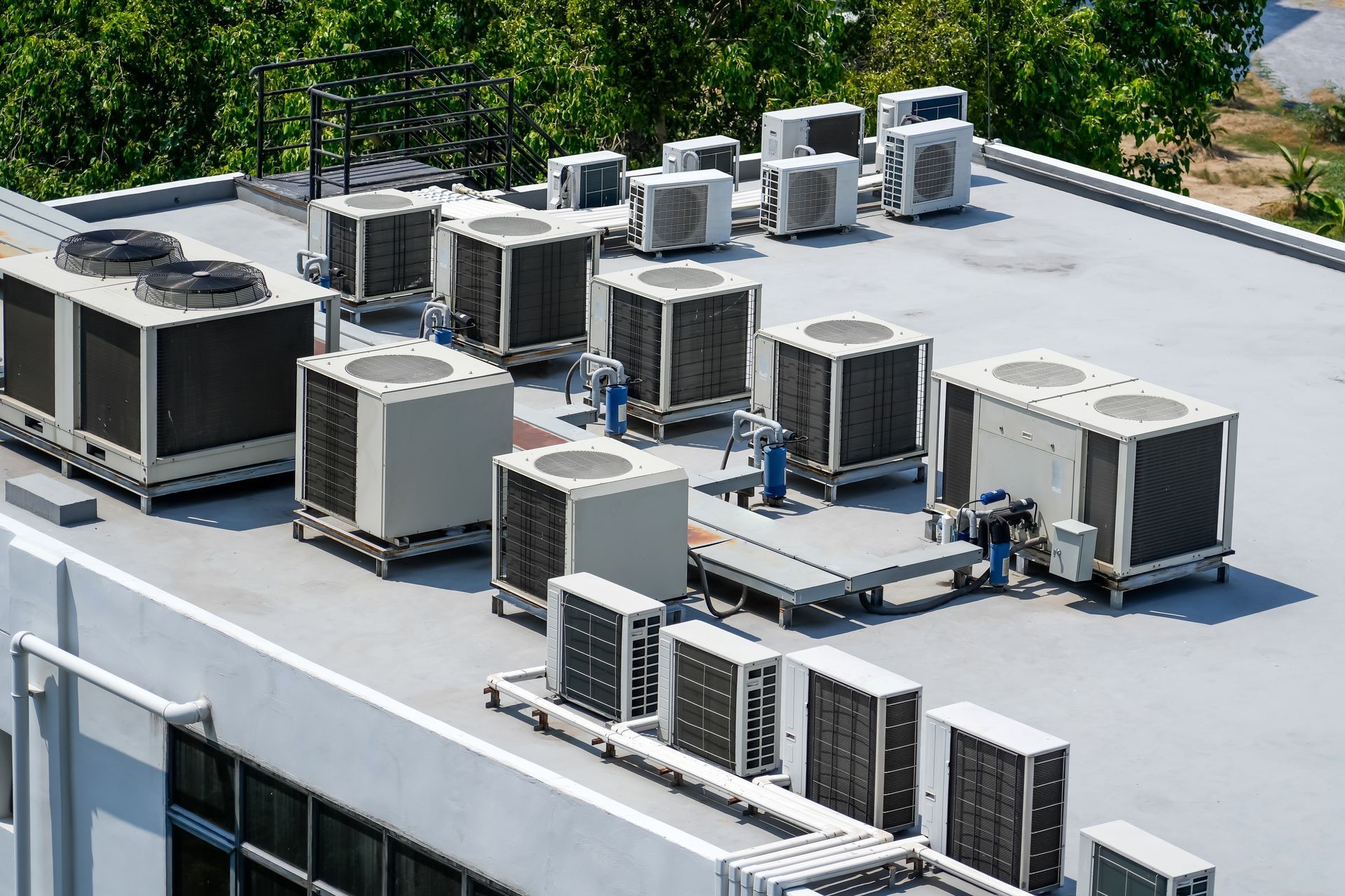 Air conditioning units on a building rooftop, grey and white, with trees in the background.