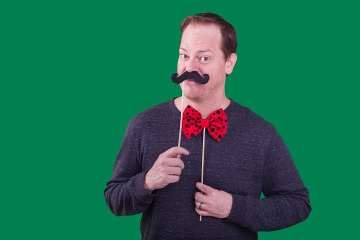 Man in dark sweater with red polka-dot bow tie and fake mustache against green background