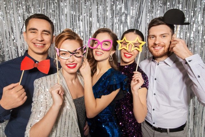Five people posing with playful props in front of a silver tinsel backdrop at a party