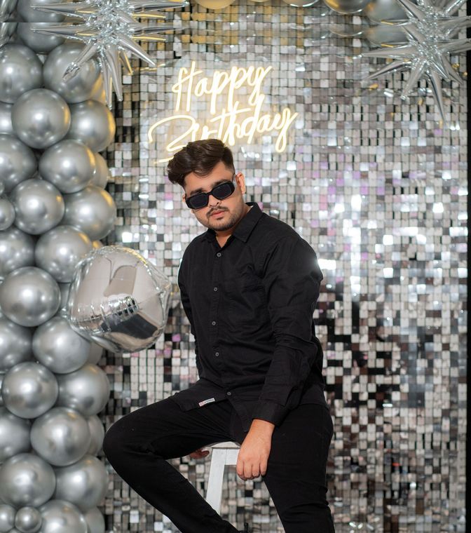 Man posing in black outfit beside silver balloons and a shimmering backdrop at a party