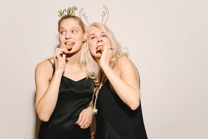 Two smiling people in black dresses wearing gold antler headbands and holding cookies at a party