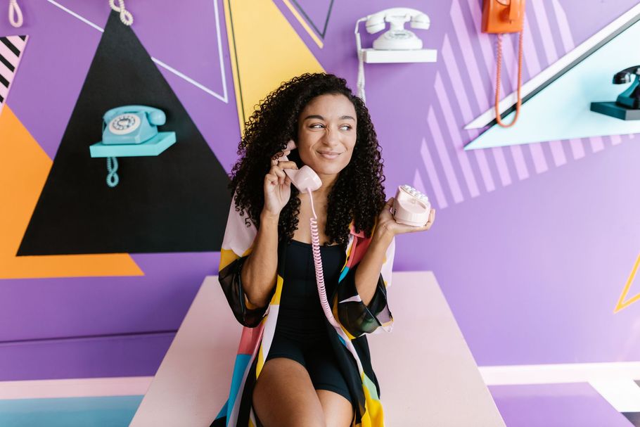 Woman smiling and holding a pink phone in a colorful retro-themed room with wall-mounted telephones