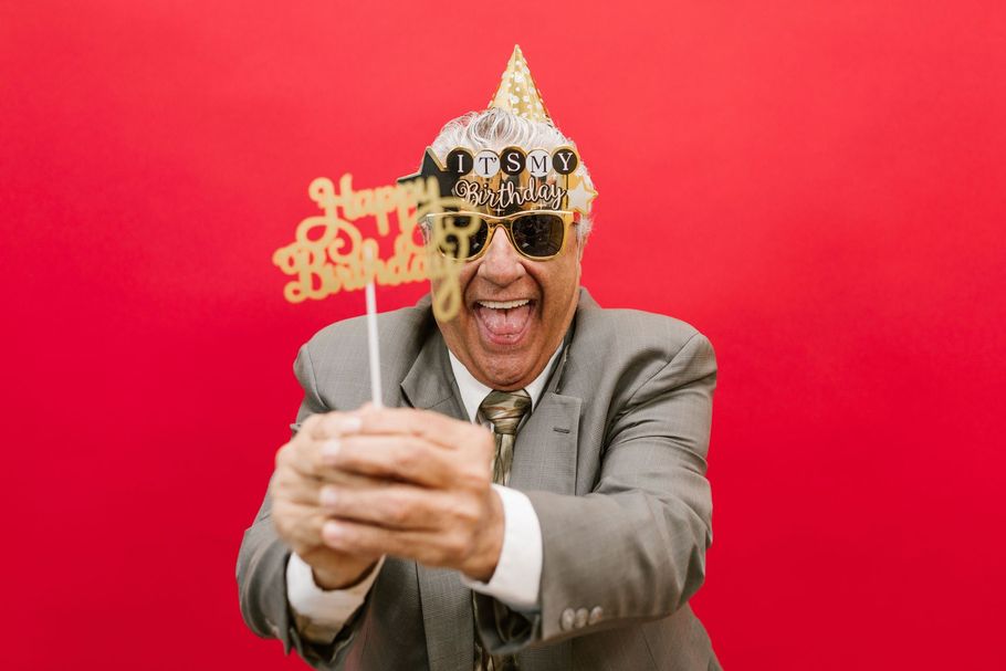 Man in gray suit and gold crown posing against a red background, holding a decorative sign and making a playful face