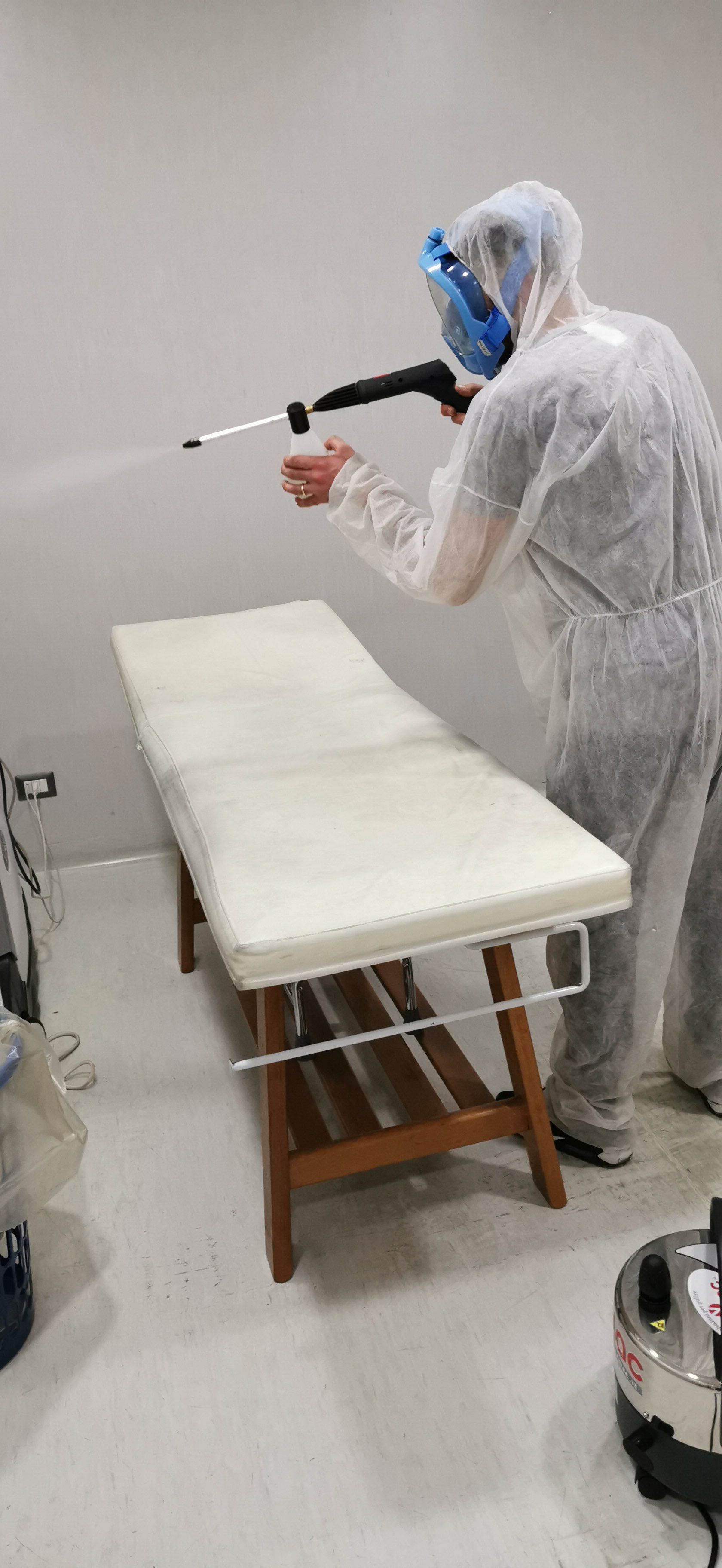 Persona in tuta protettiva che spruzza disinfettante su un lettino da massaggio in una stanza.