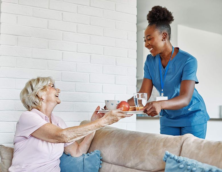 Caregiver in blue scrubs hands an apple and drink to a smiling woman on a couch.