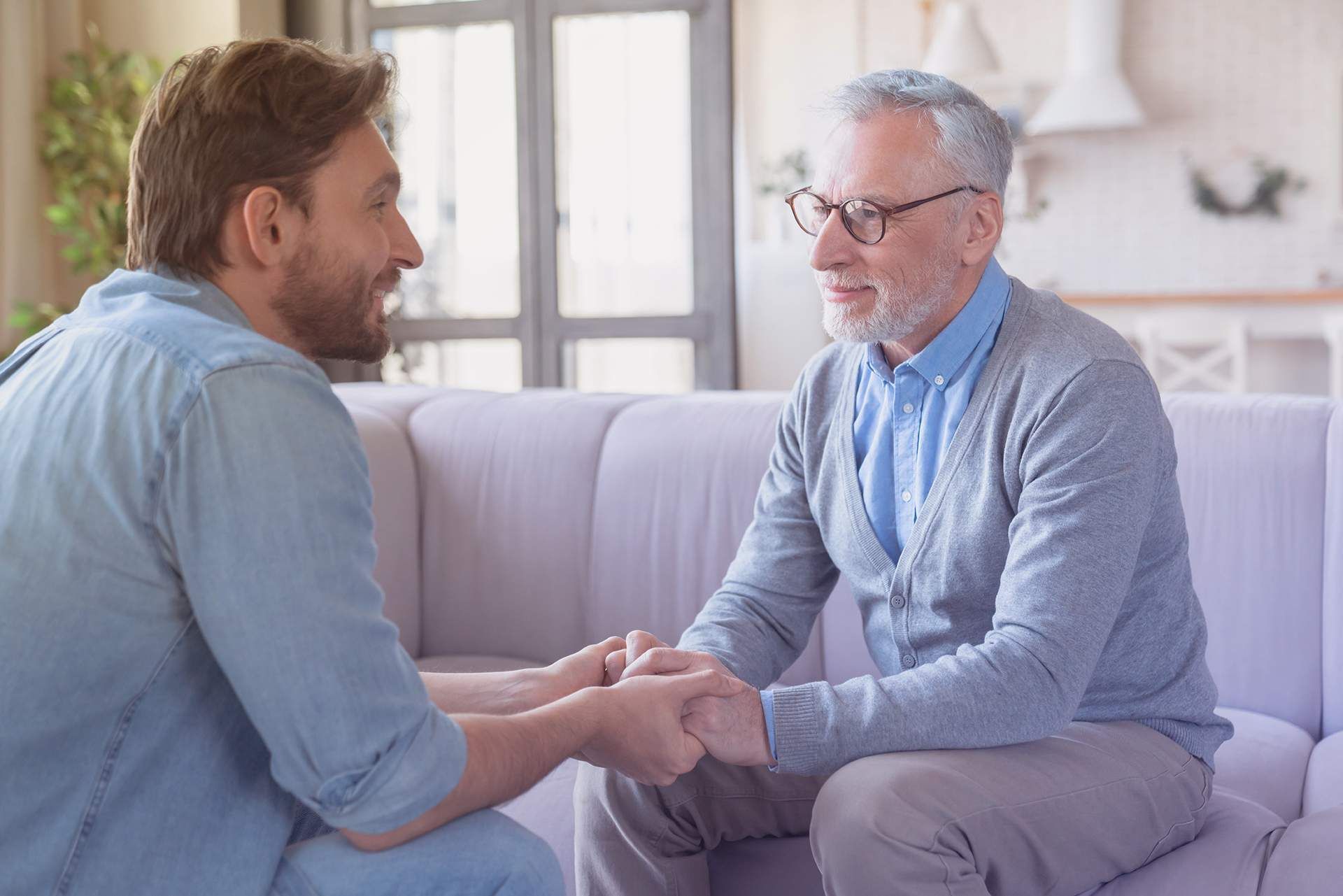 Two men sit on a couch, holding hands and talking in a bright living room.