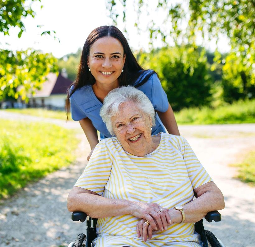 Woman pushing smiling elderly person in wheelchair on a sunny path outdoors