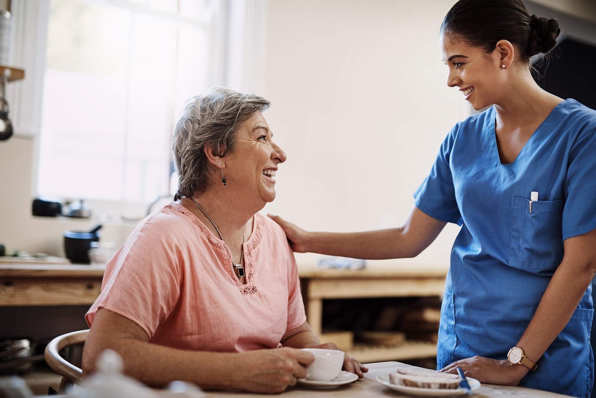 Caregiver in blue scrubs comforts a smiling older woman at a table with coffee in a bright home setting