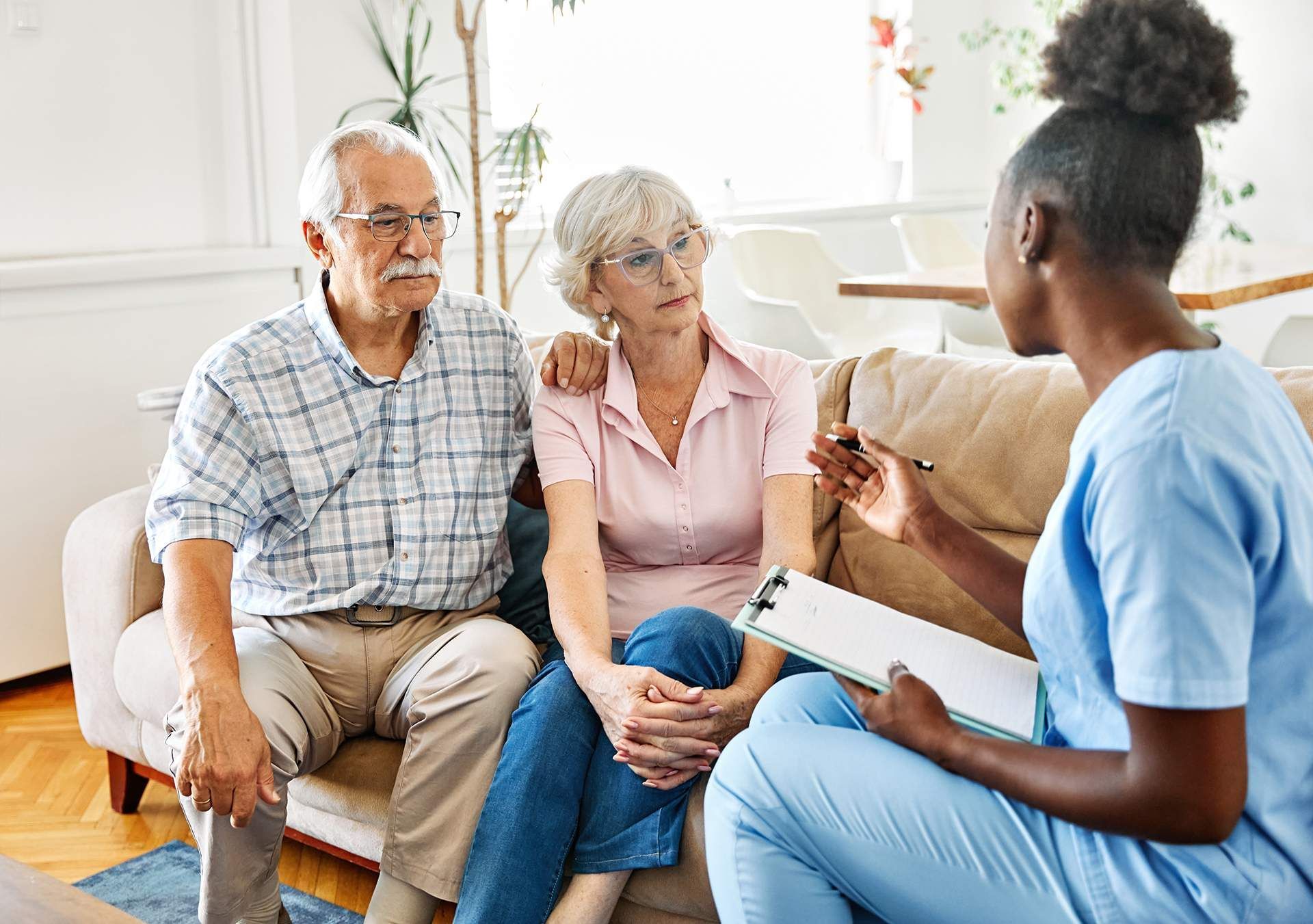 Two people listening to a nurse during a home visit, seated on a couch in a bright living room