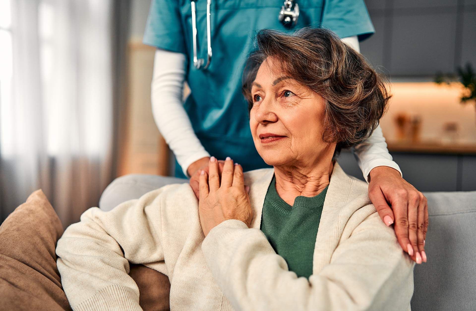 Elderly woman sitting with a caregiver’s hand on her shoulder in a cozy indoor care setting