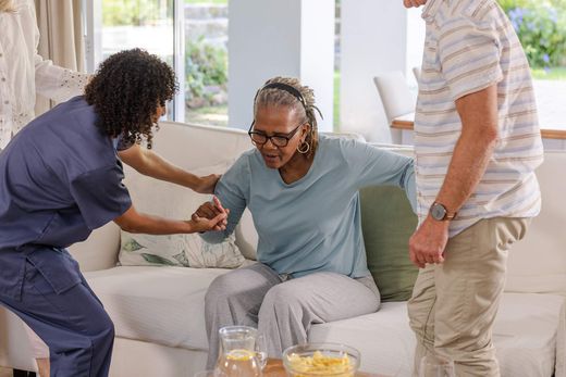 Caregiver helping a distressed person sit up on a couch while another person stands nearby in a bright living room