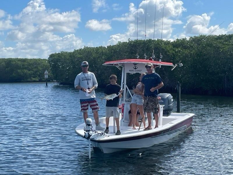 Four people on a fishing boat in water, holding fish. Sunny day with trees in background.