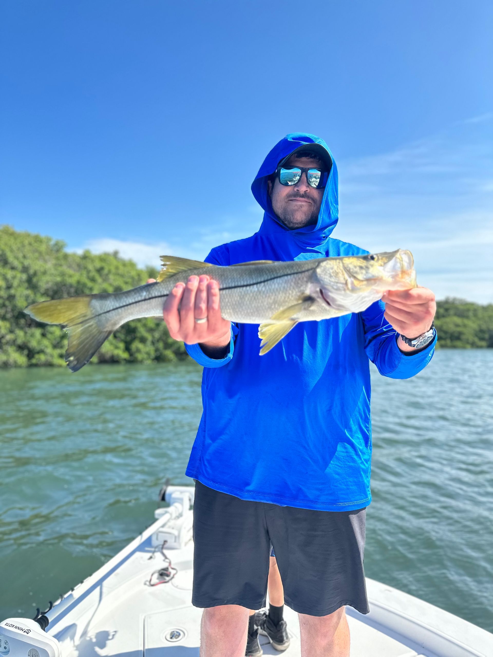Man on boat holding up a snook fish, wearing blue hoodie, sunglasses, and black shorts, against a blue sky.