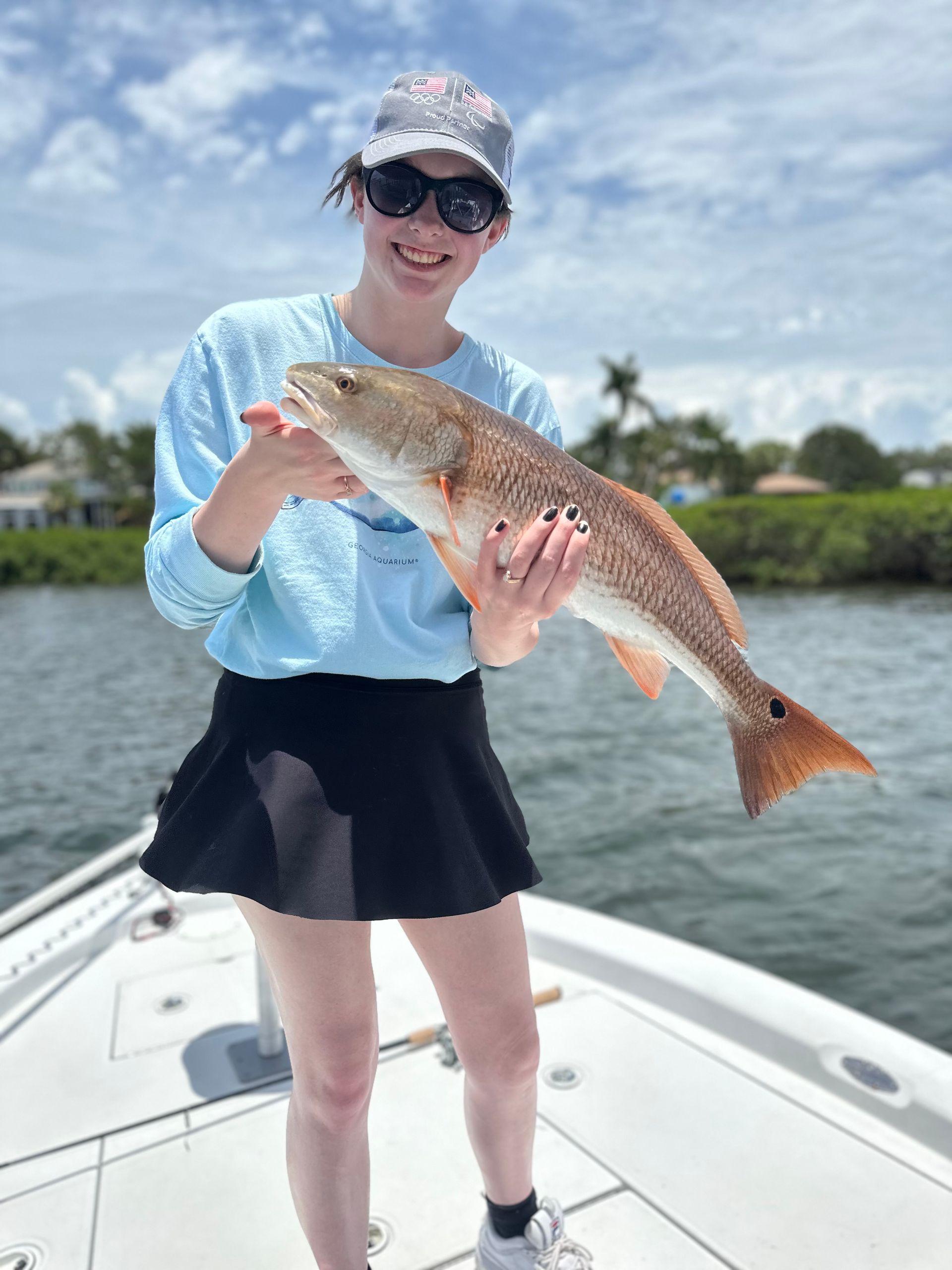 Young person on a boat holding a reddish-brown fish. Bright sky and water.