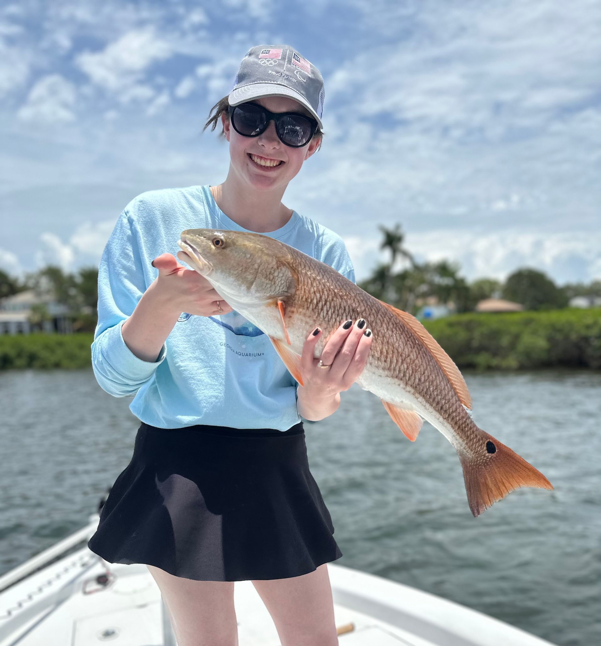 Person on a boat holding a reddish fish. Sunny day, wearing hat and sunglasses, smiling.