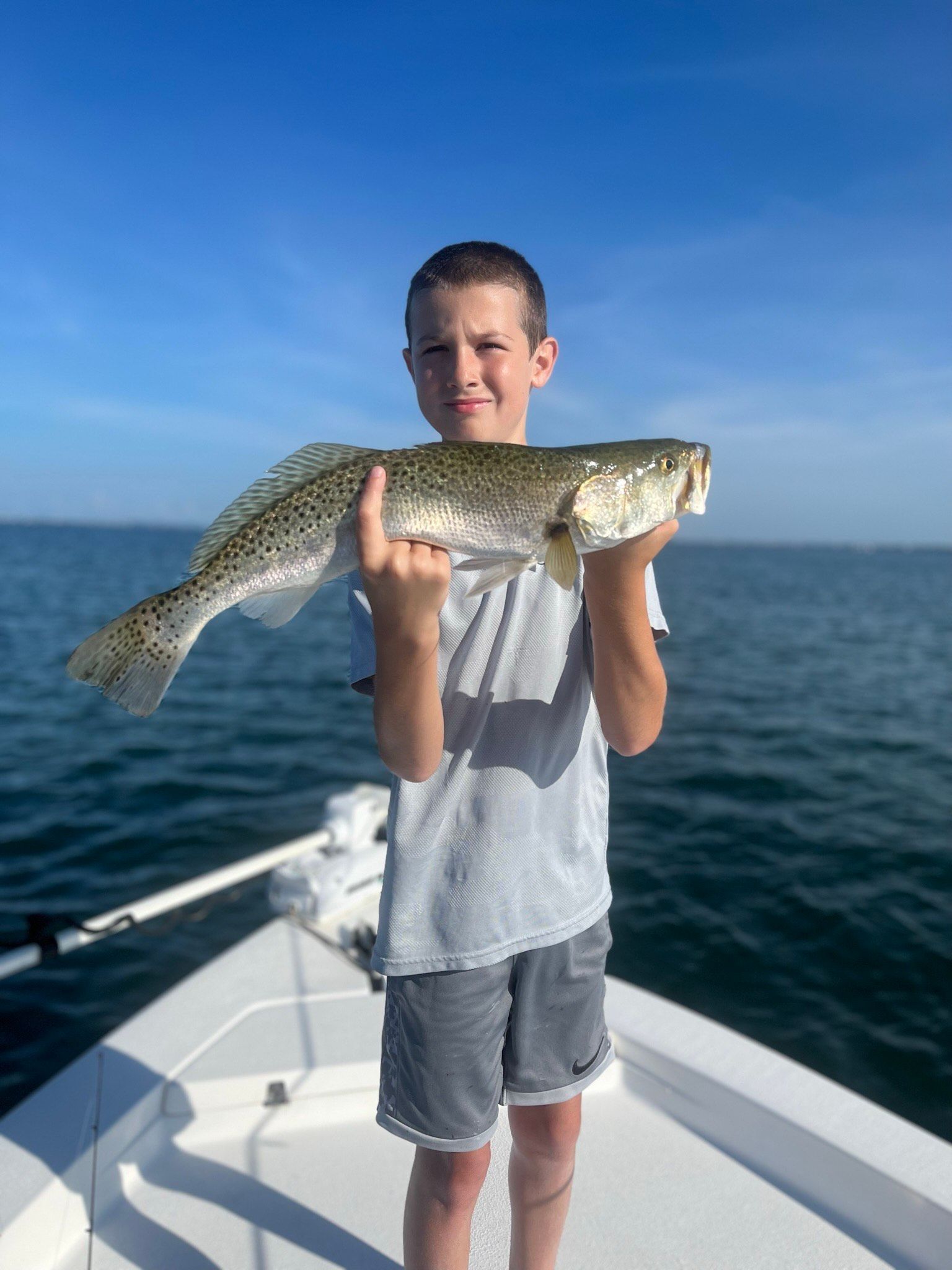 Boy on a boat holds up a spotted speckled trout against a blue sky.