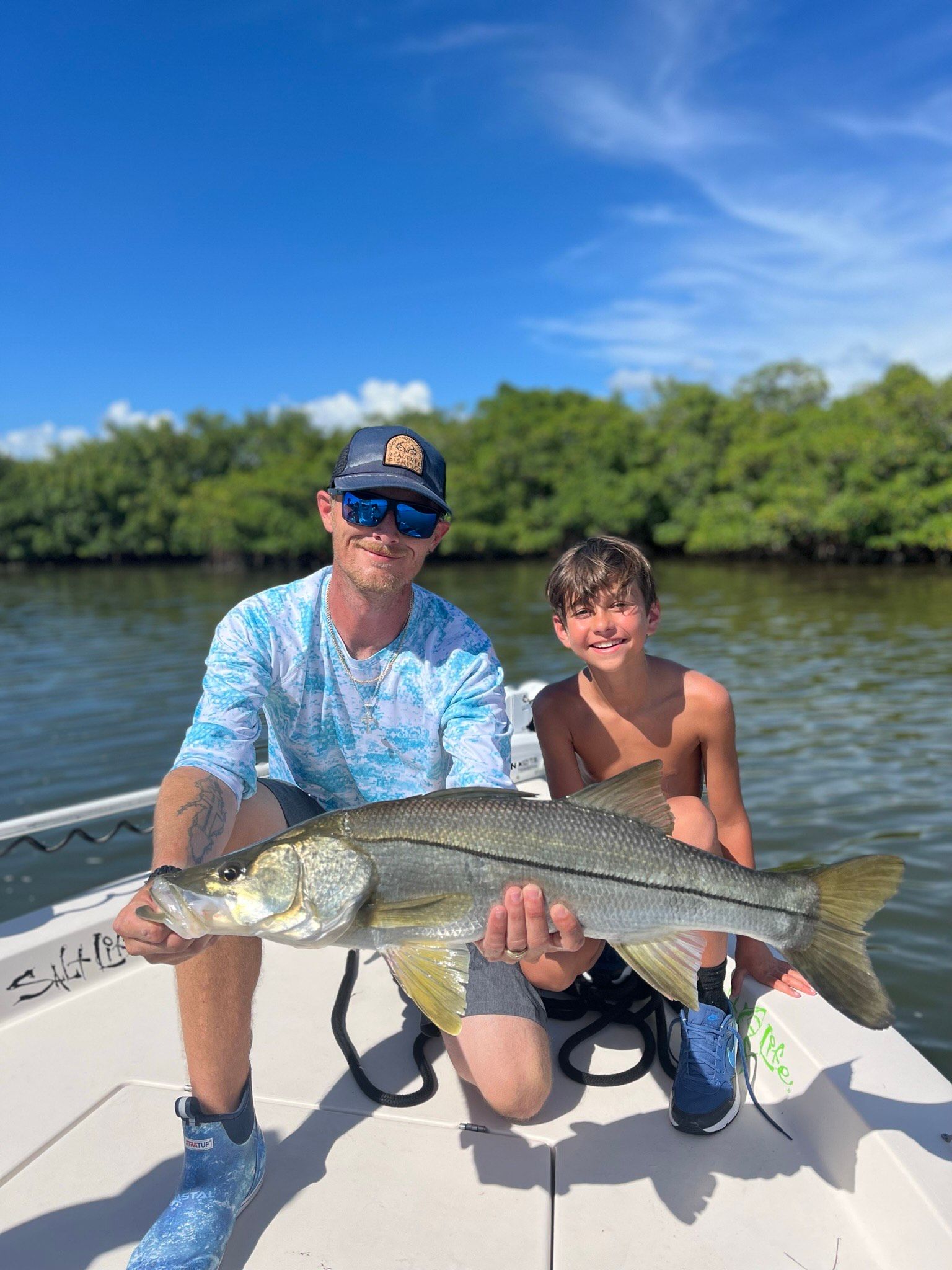 Man and boy on a boat hold a large fish. They are smiling in a sunny, green-water setting.