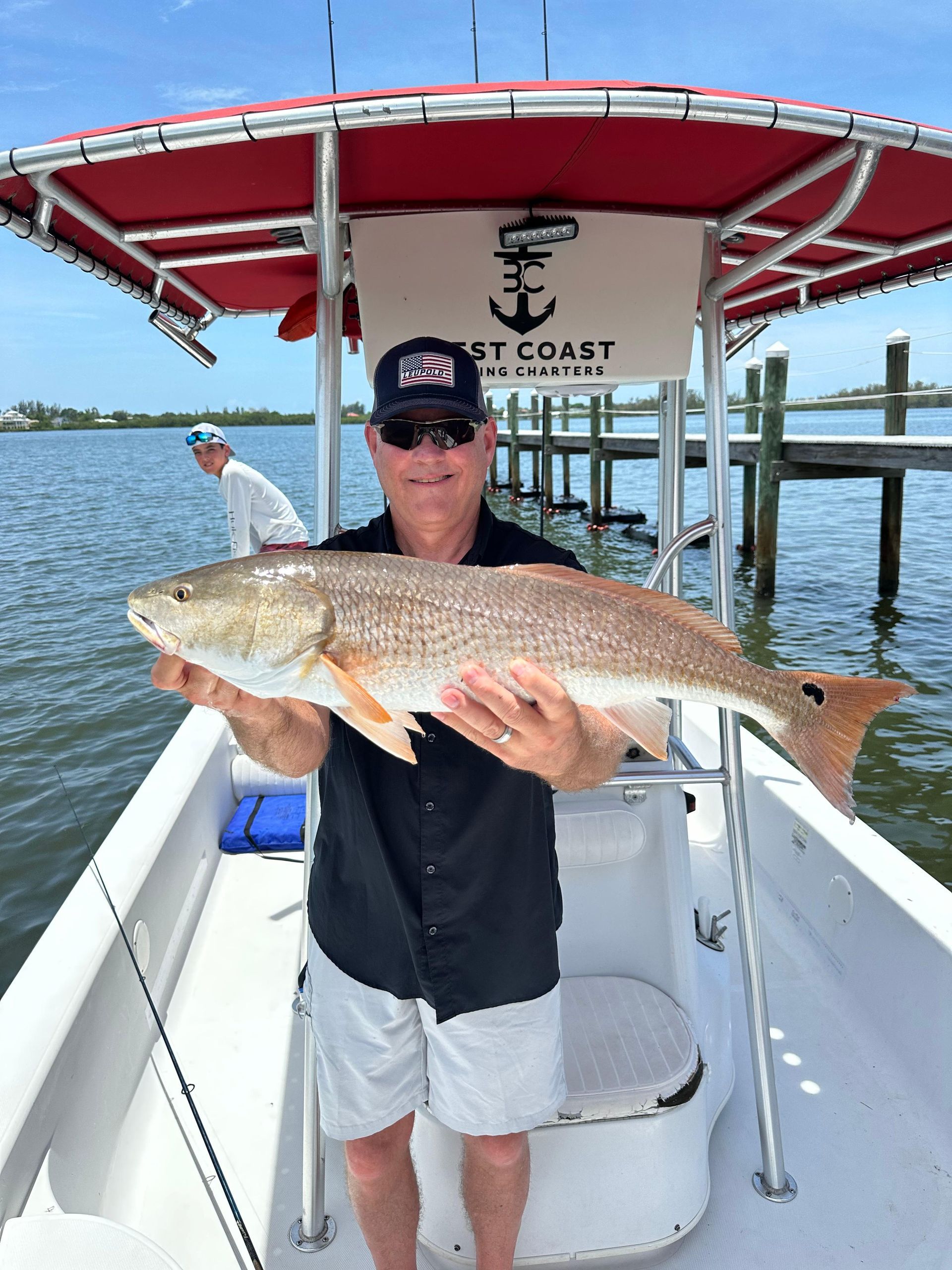 Man on boat holds a large redfish. Bright sun, water and dock in the background.