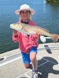 Young person kneels, holding a large fish. They wear a hat, pink shirt, blue shorts, and are on a boat with water in the background.