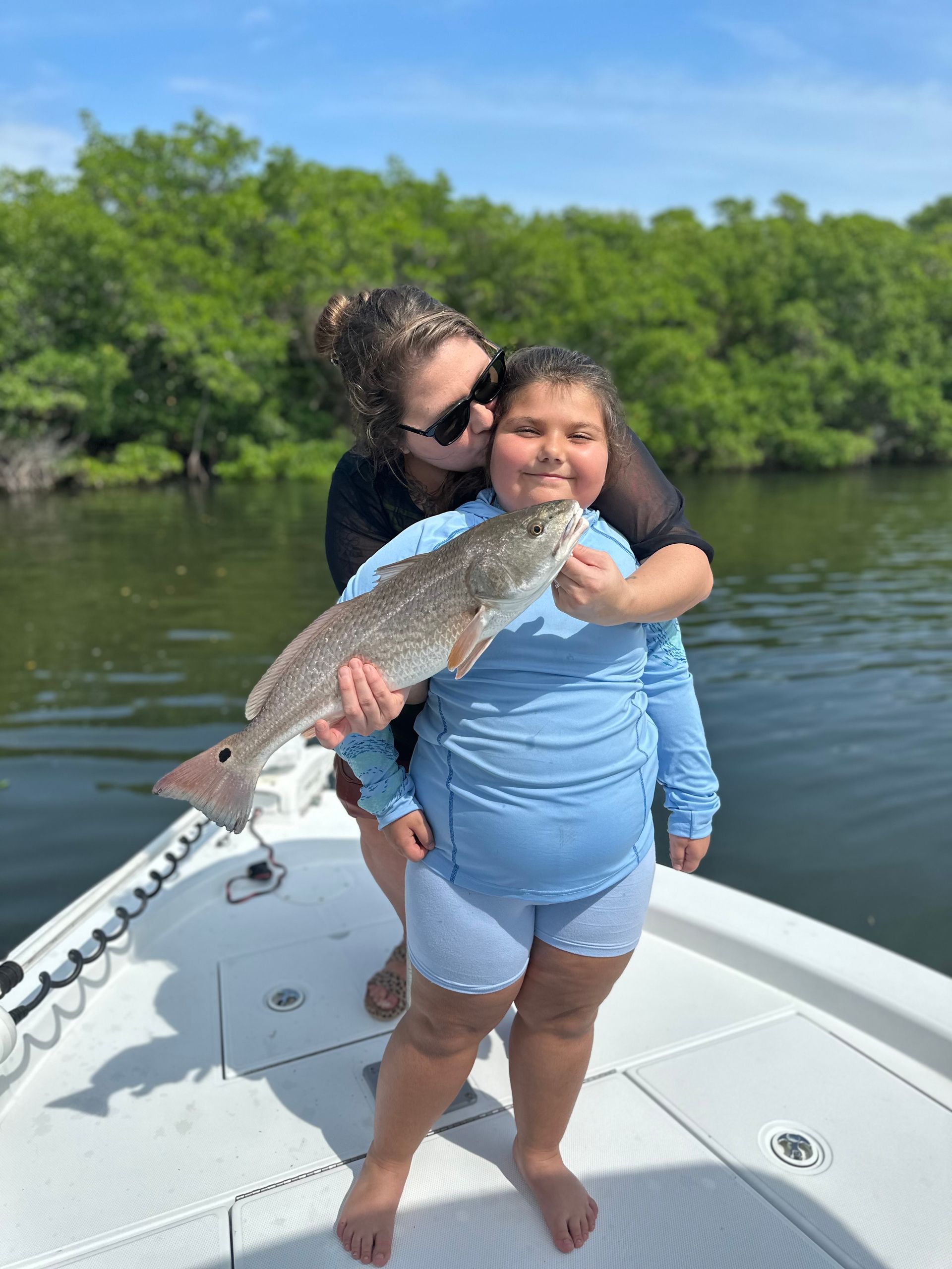Woman and child on boat holding a large red fish, woman kissing child's cheek, green trees in background, sunny day.