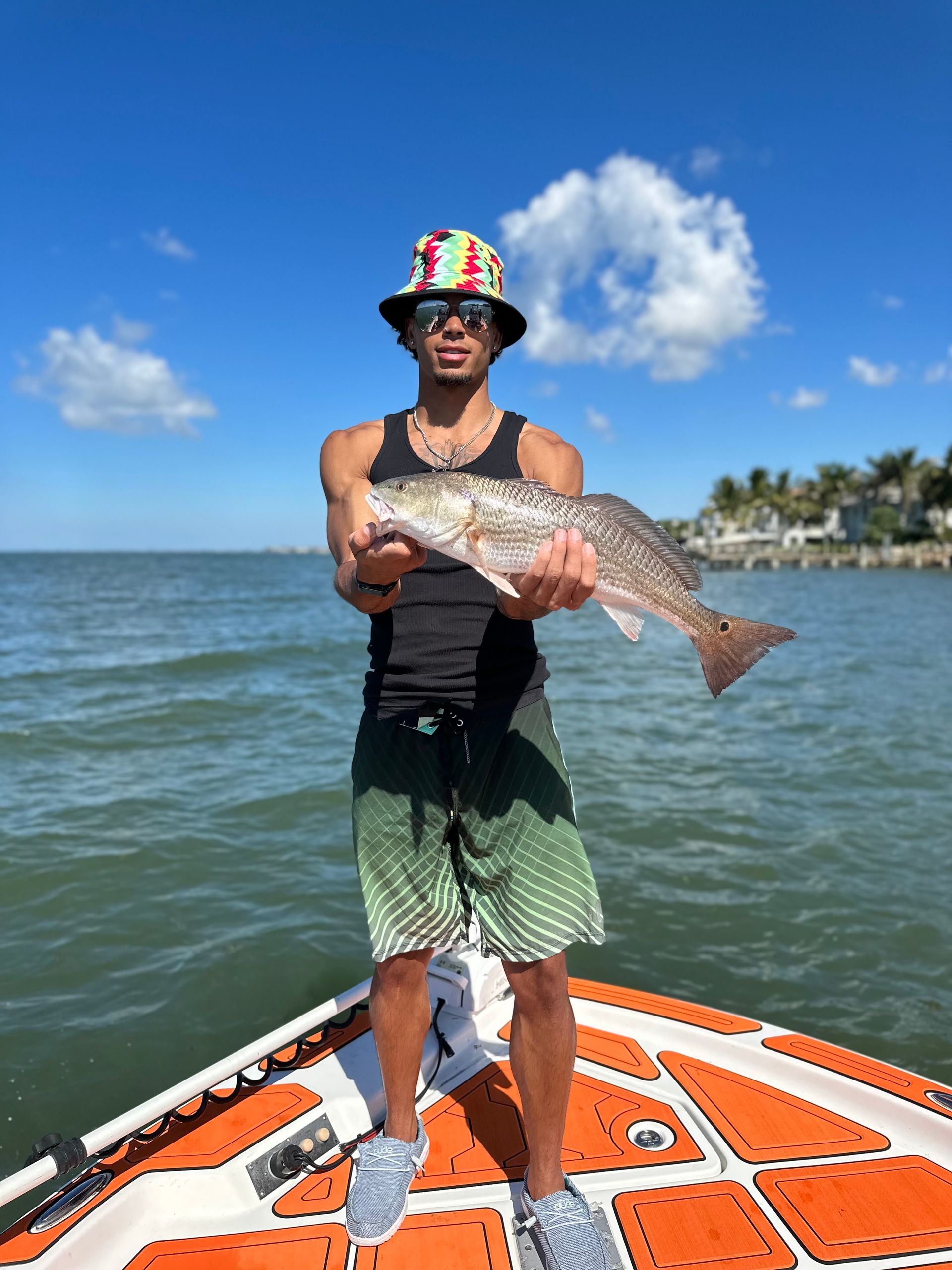 Man on boat holds up a fish, blue sky, water, colorful bucket hat.