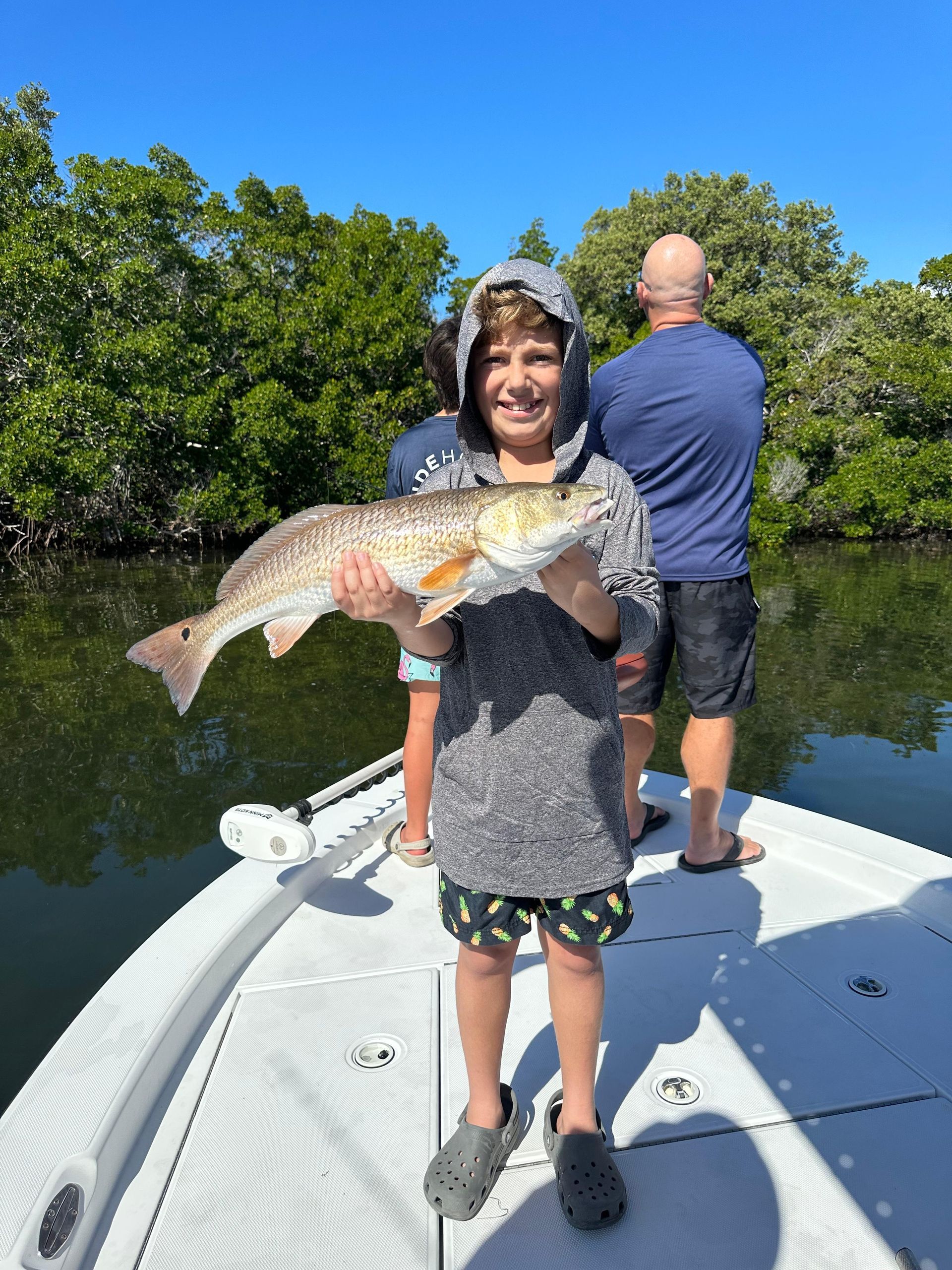 Boy smiles, holding large red fish on boat, sunny day, another man in background.