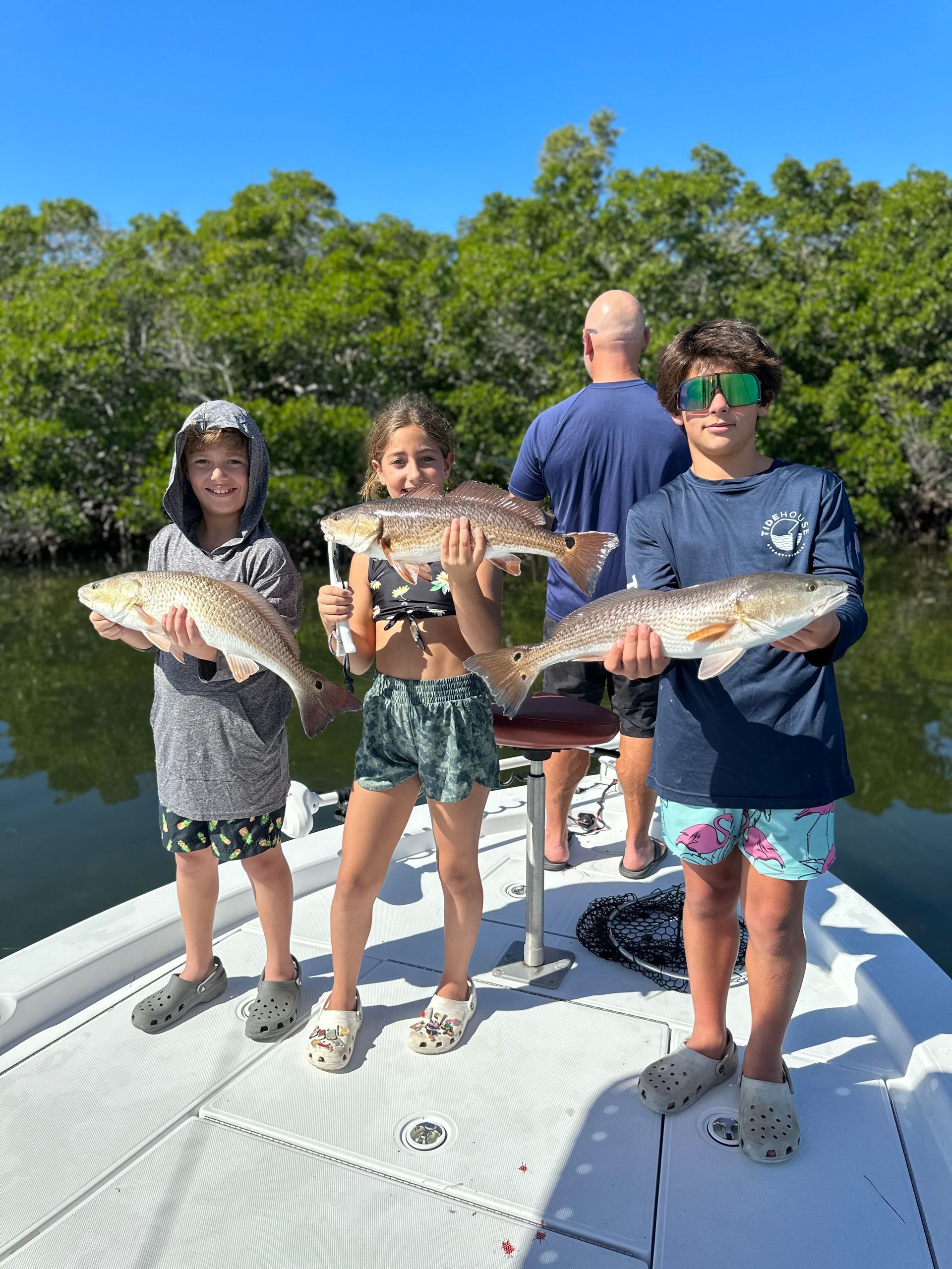 Three children on a boat hold up fish they caught; an adult stands behind them. Sunny day, green trees.