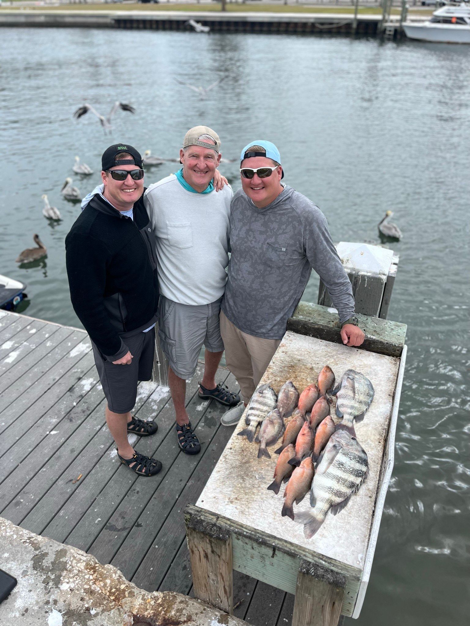 Three men posing on a pier with a fish cleaning table and a pile of fish. Water and pelicans are in the background.