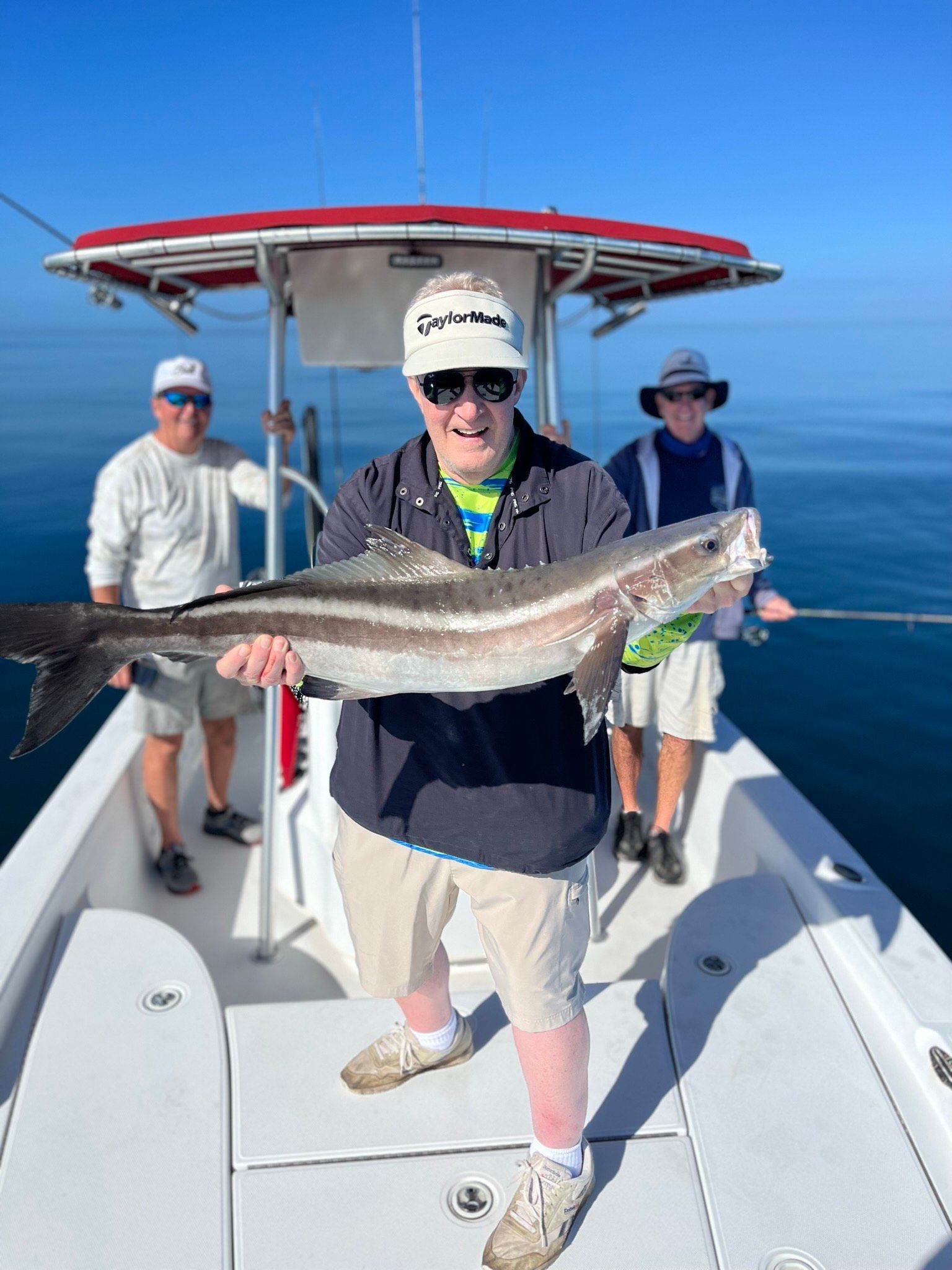 Man on a boat holding a large fish, two others behind him. Blue water, sunny day.
