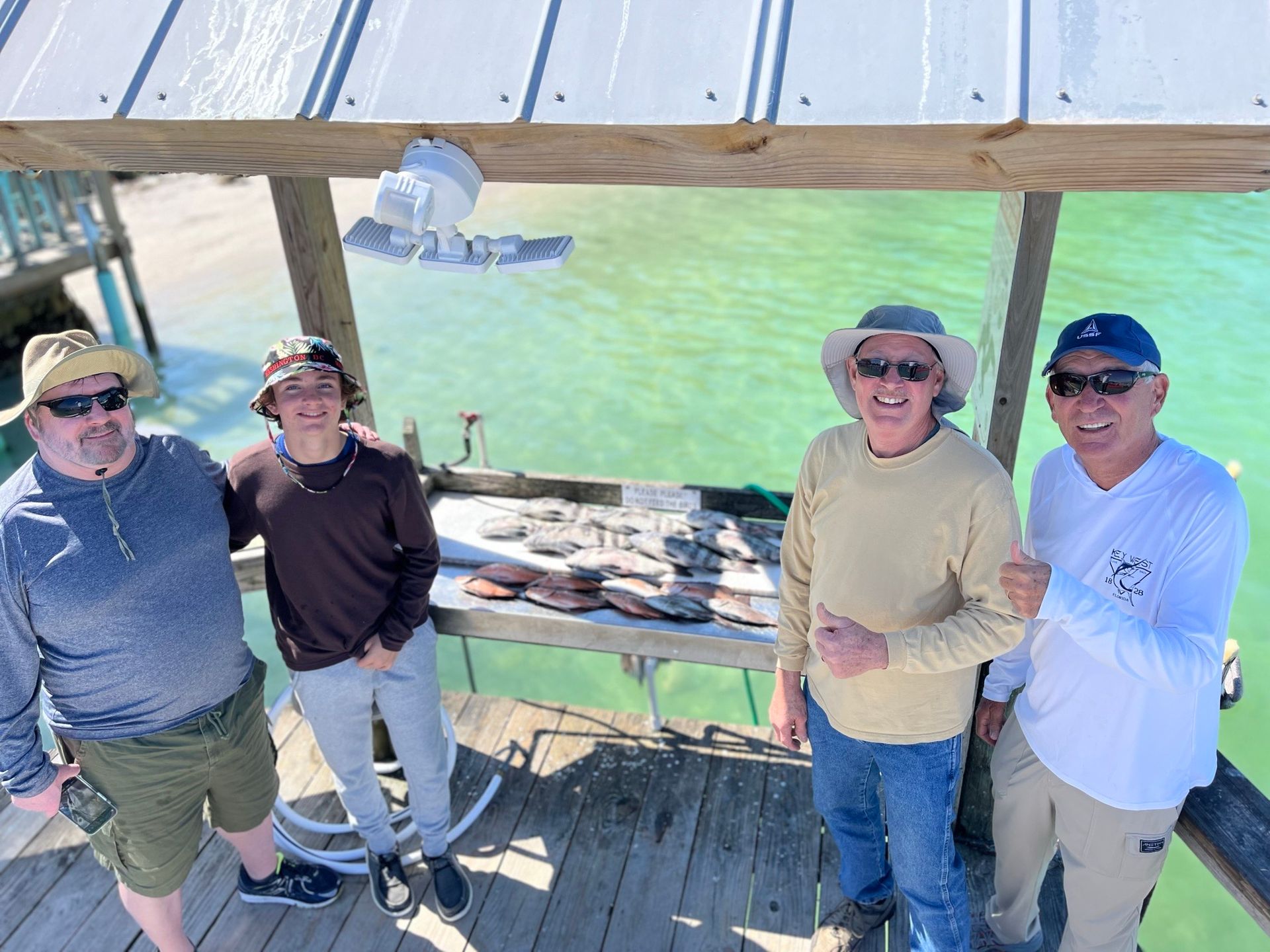 Four people standing on a pier, smiling. A table displays fish. Clear turquoise water in the background. Sunny day.