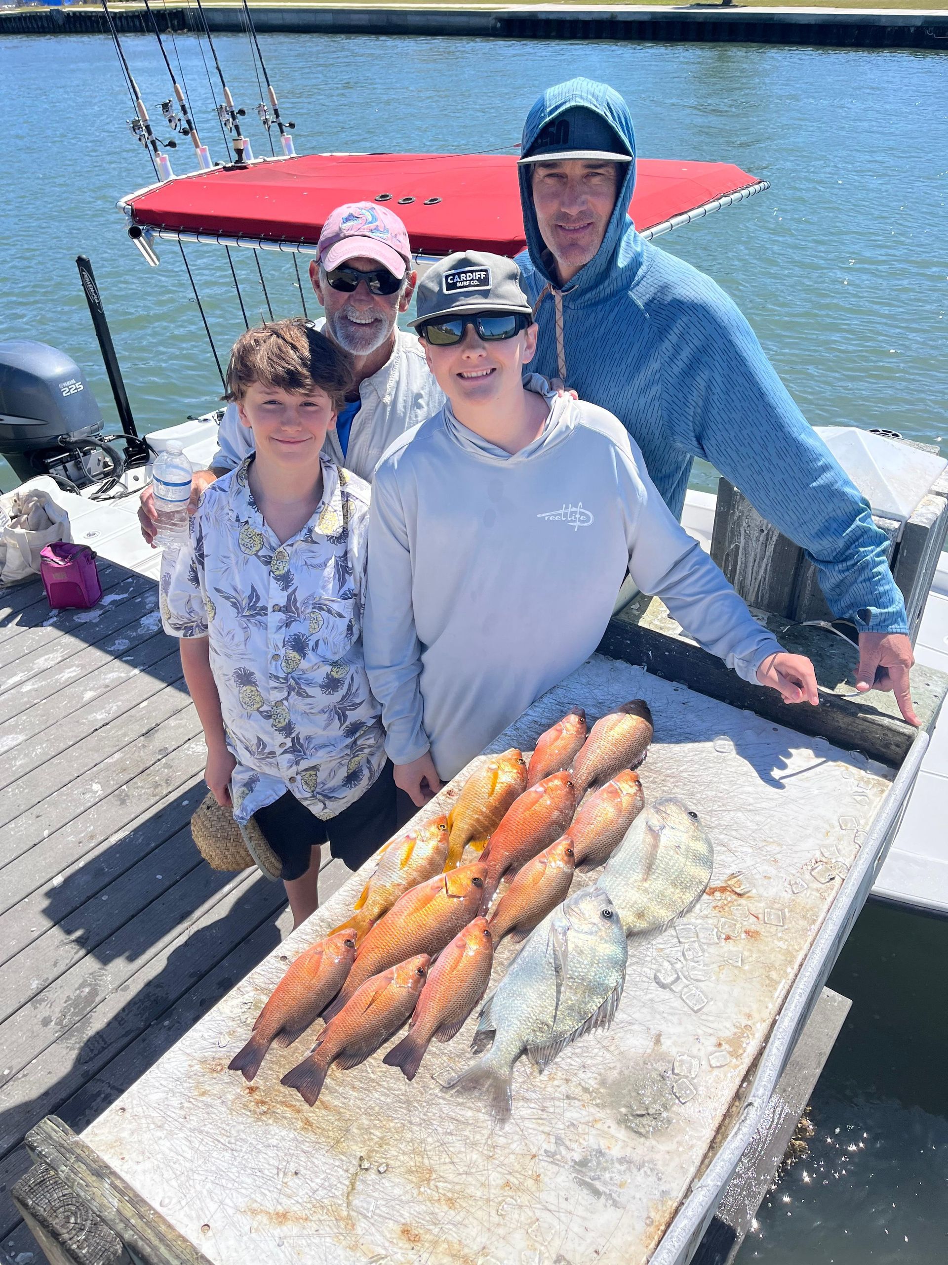 Family on a boat dock displays freshly caught red fish. Sunny day; man points, others smile.