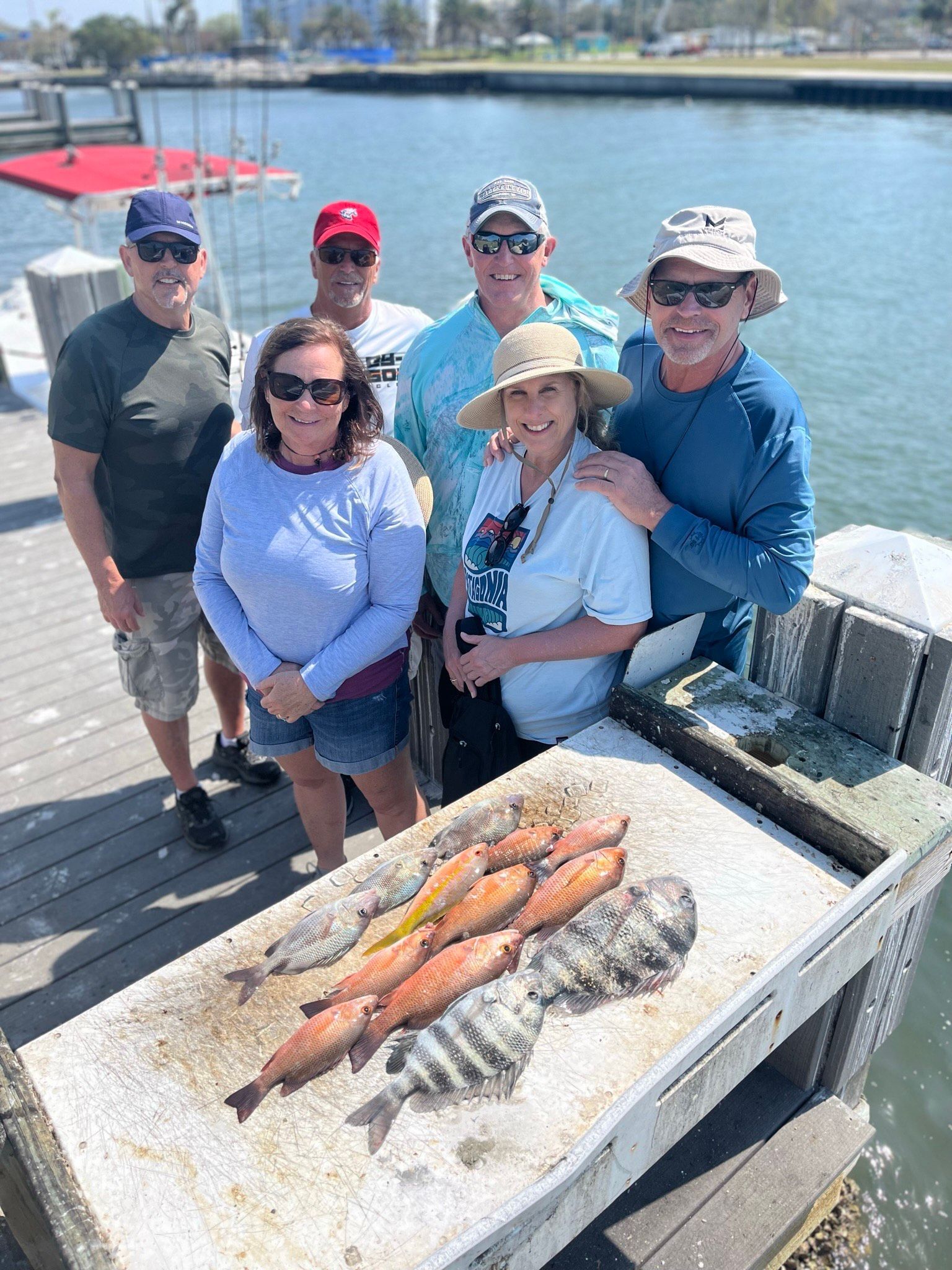 Group of people pose with fish on a pier; sunny day with water and buildings in the background.