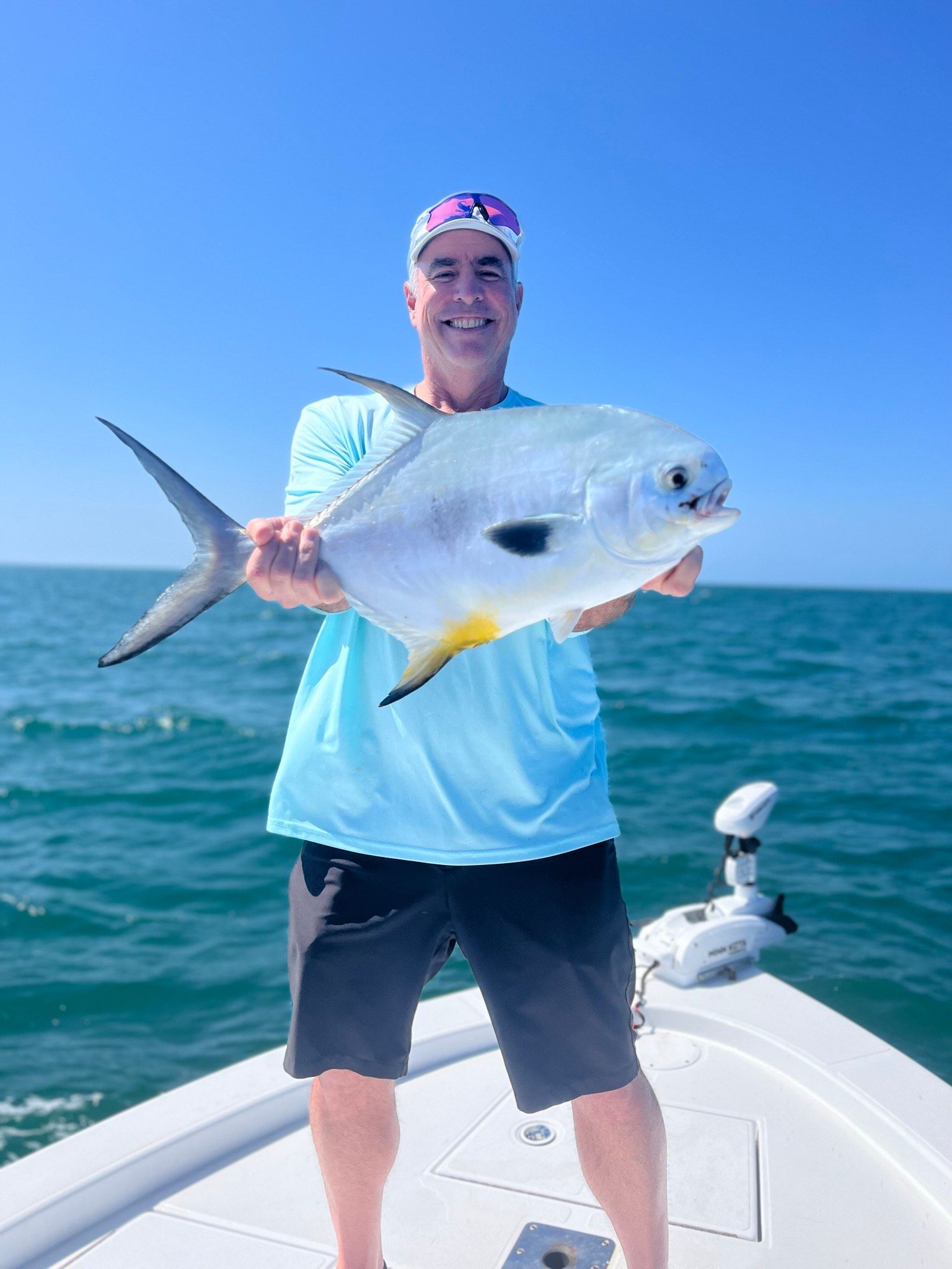 Man holding a large, silvery fish on a boat. Ocean, blue sky. Fish has a yellow fin.
