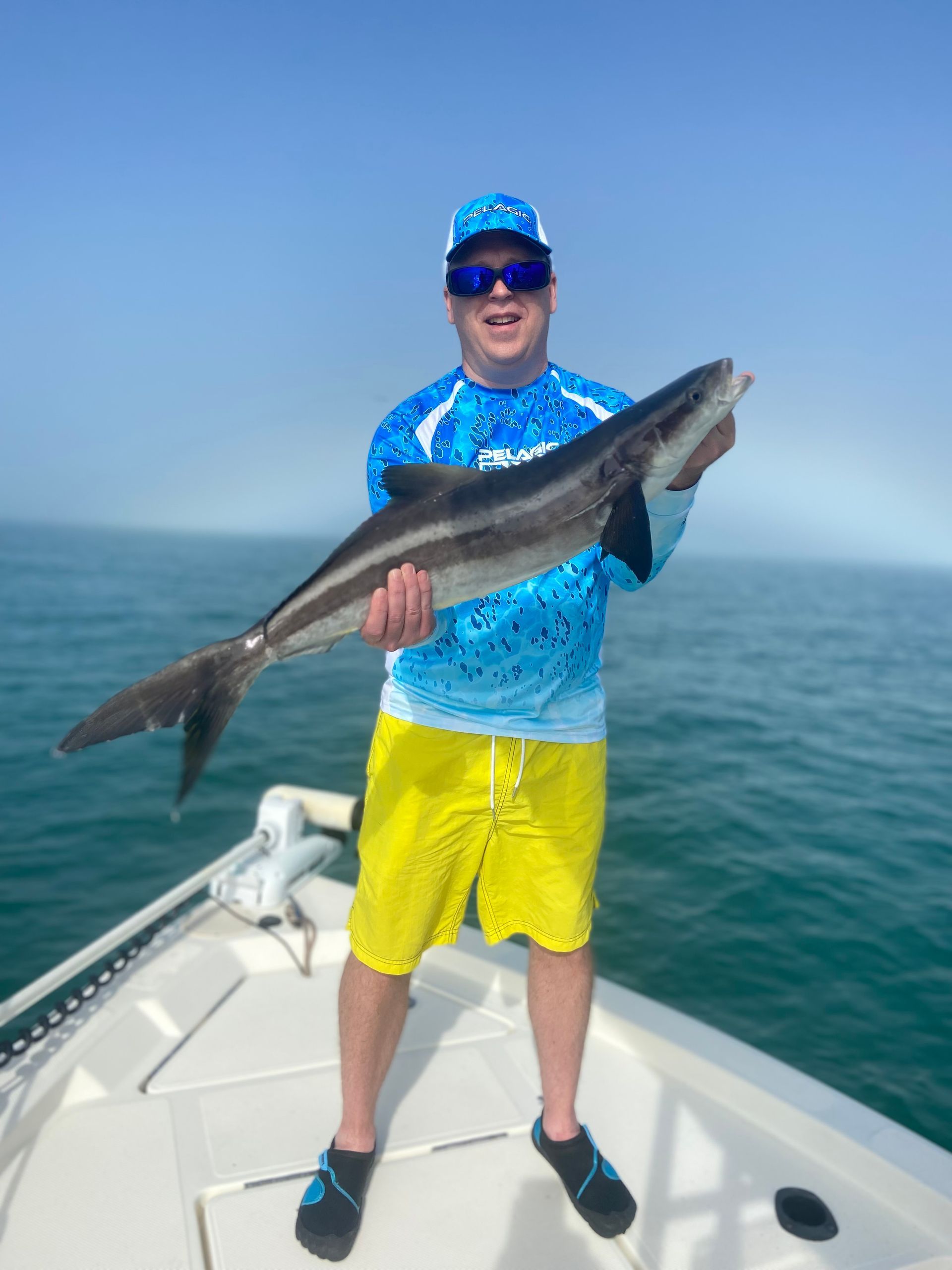 Man on boat holding a large fish, blue water and sky in background.