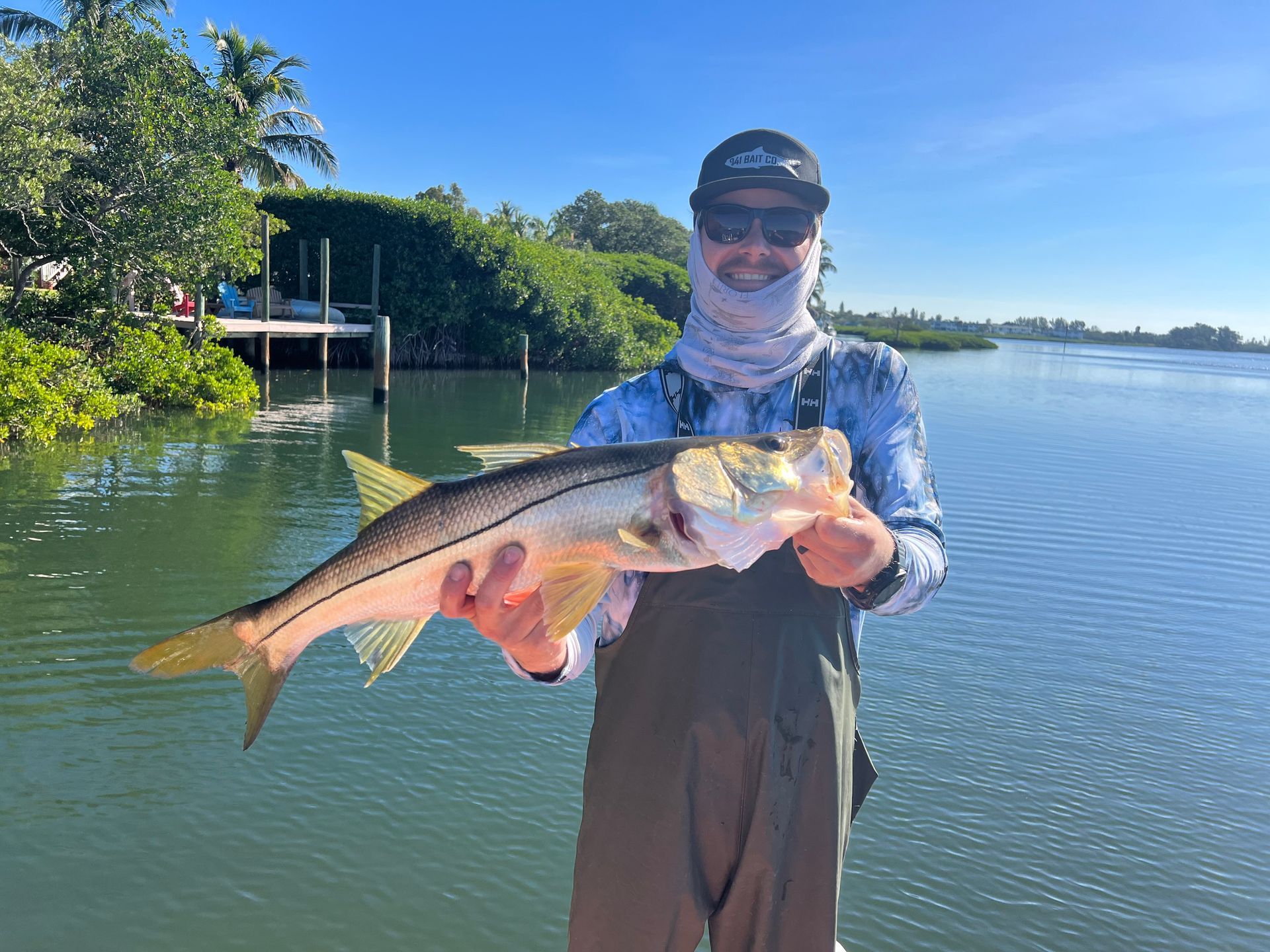 Man in waders holds a large, silver snook fish on a dock, water and greenery in background.