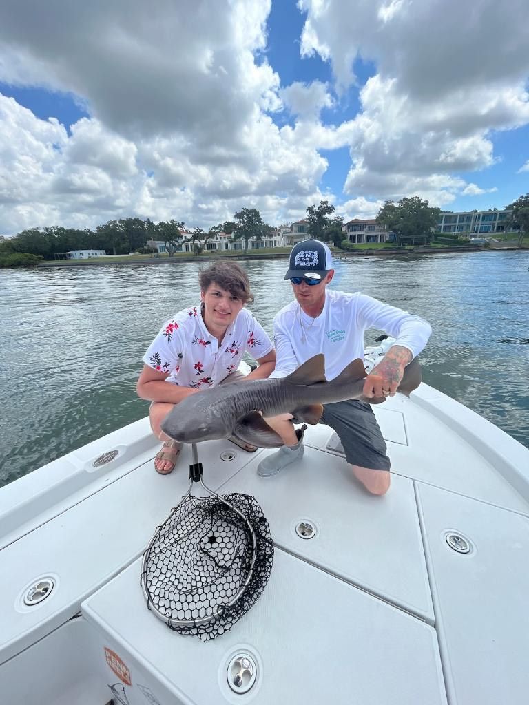 Two people on a boat with a fish. One holds the fish, the other smiles. Cloudy day, water and houses in background.