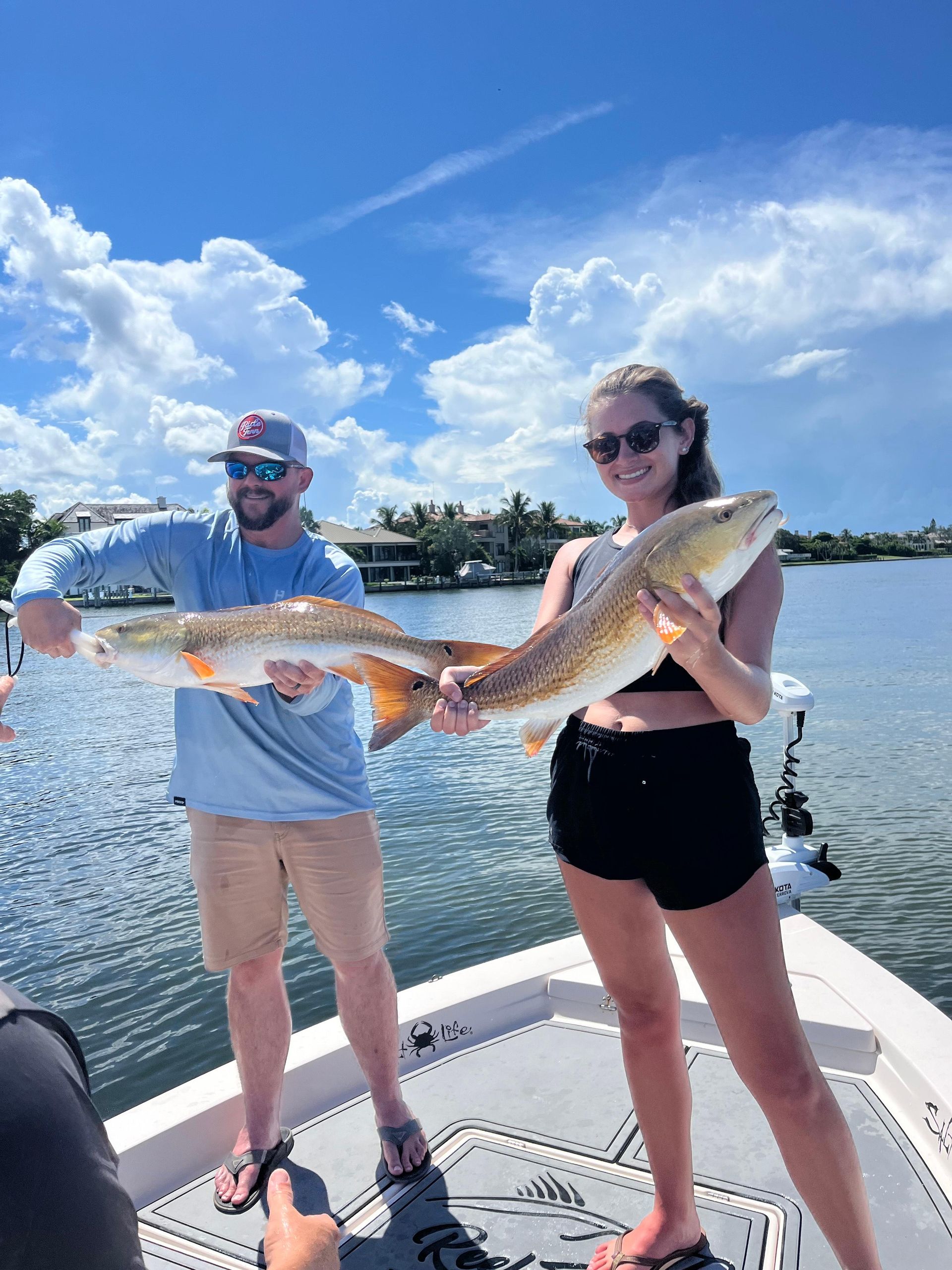 Man and woman on a boat holding a large, reddish-orange fish. Blue sky and water in the background.