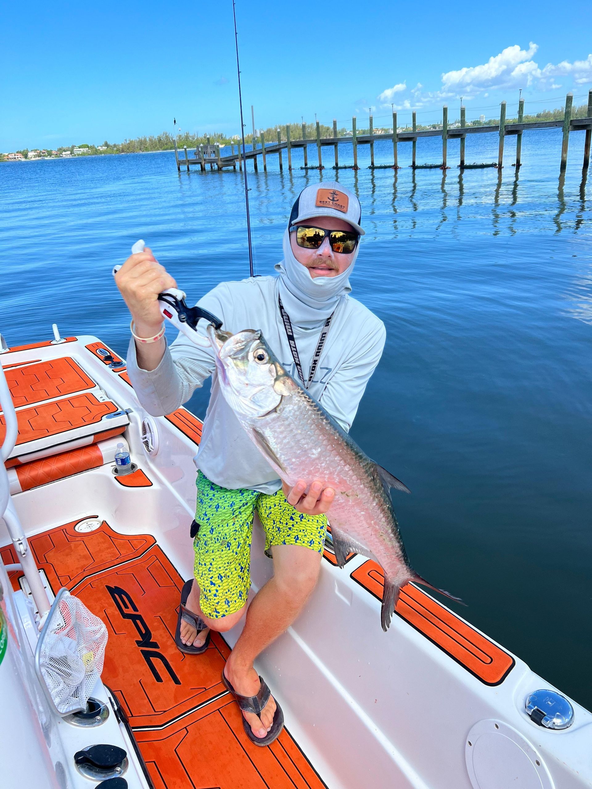 Person on a boat holding a large fish, sunny day, water and pier visible.