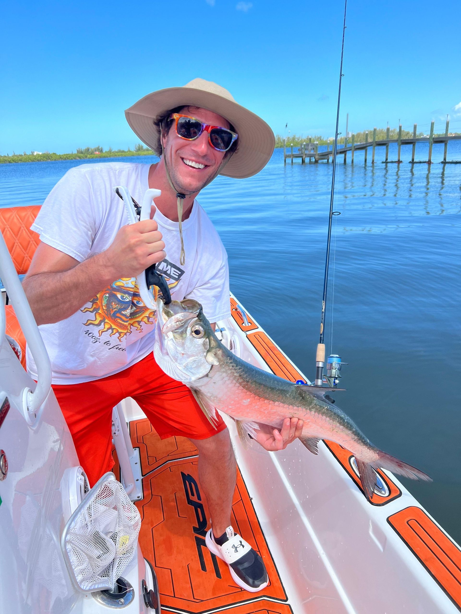 Man on boat smiles, holding a caught fish with pliers; bright blue water and sky.