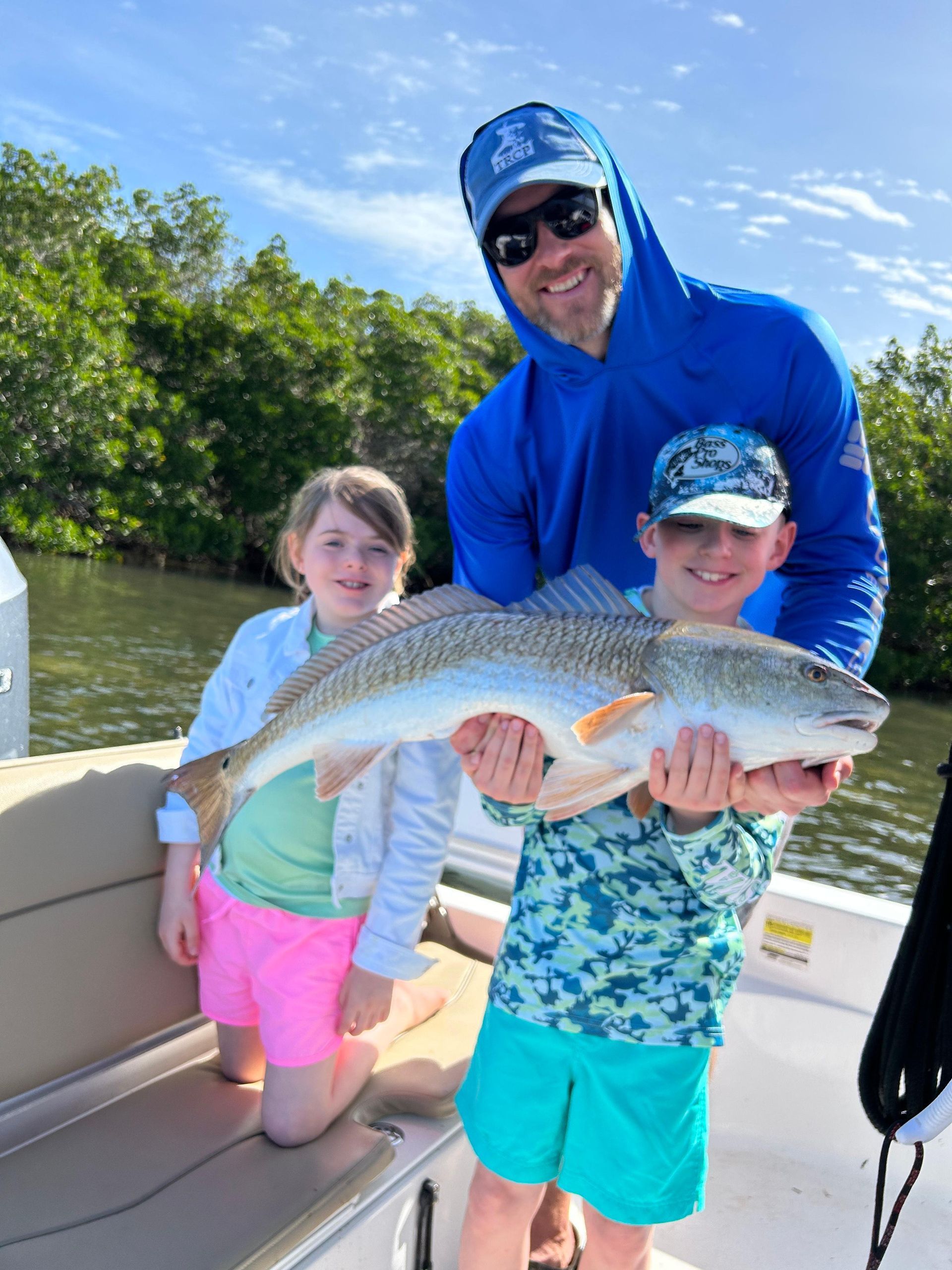 Man and two children on a boat holding a large redfish. All are smiling, in bright clothes, near mangroves.