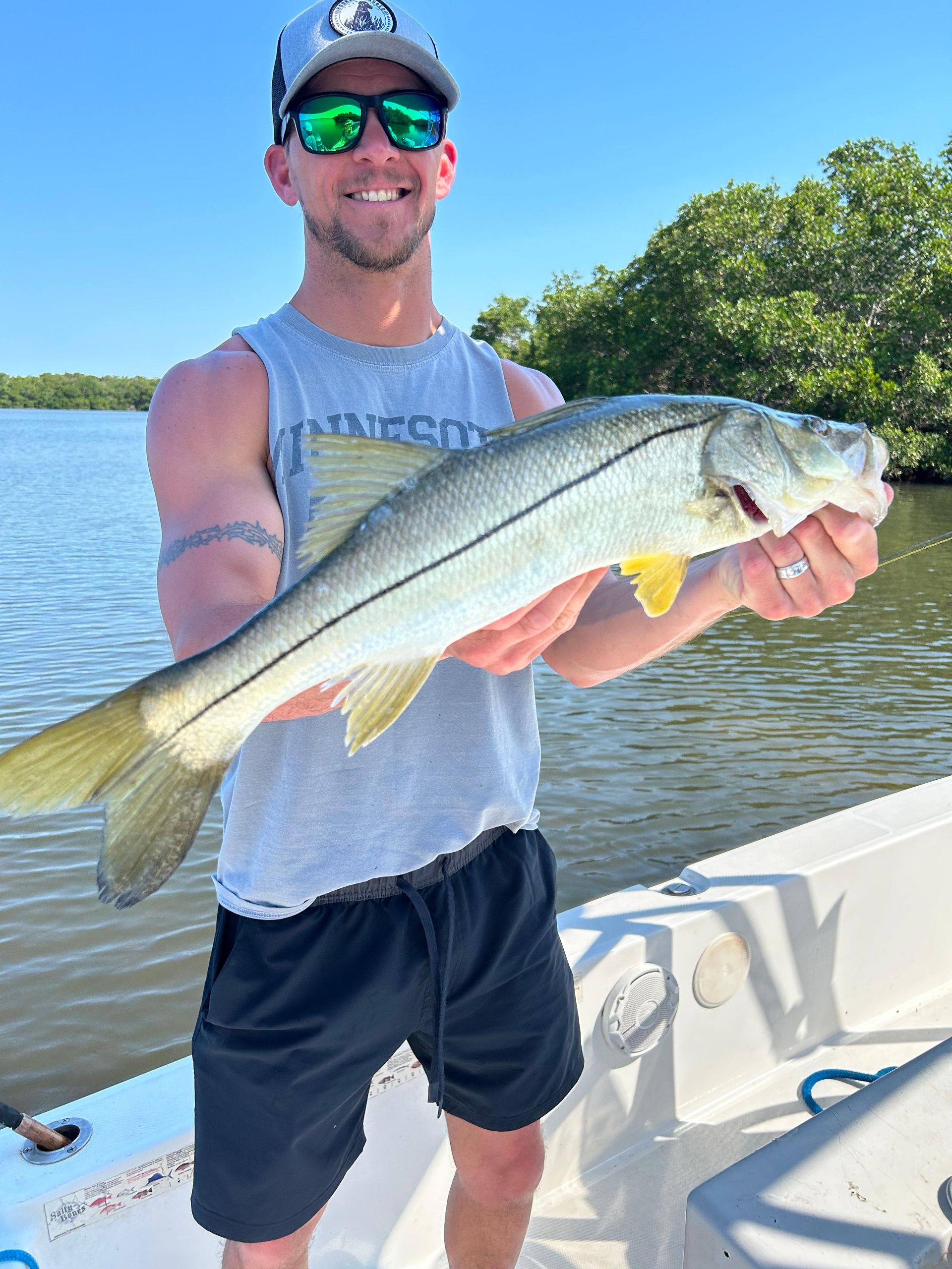 Man holding a Snook fish on a boat. He wears a hat, sunglasses, and smiling. Bright day with water and mangroves.