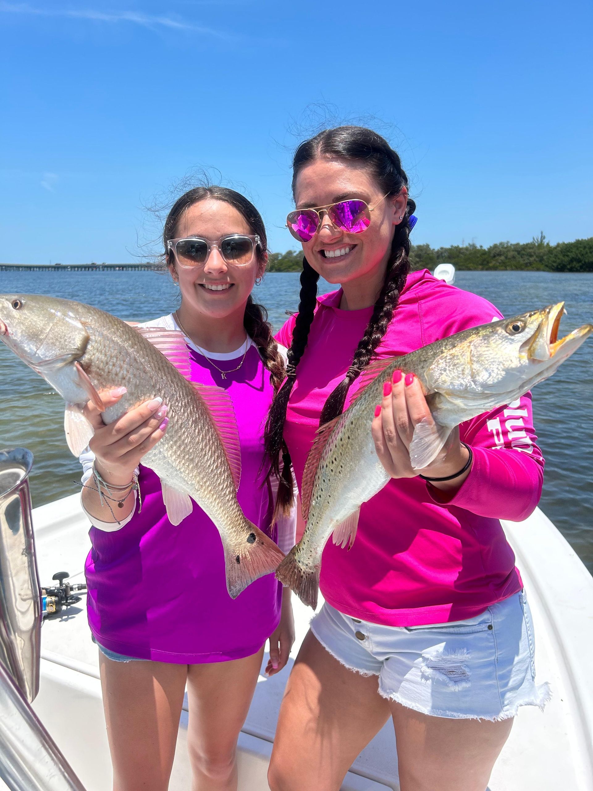 Two people on a boat holding fish. They wear pink shirts and sunglasses. Bright sky and water in the background.