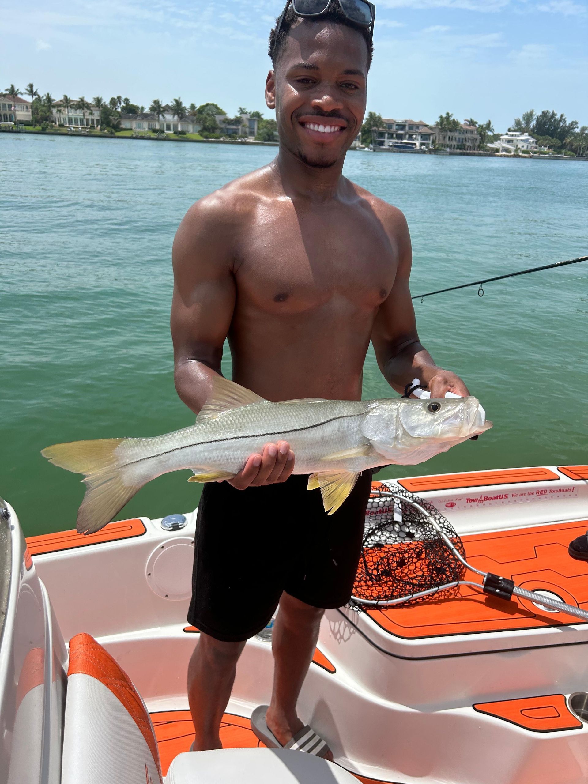 Man on a boat, holding a fish, smiling. Green water, houses in the background.