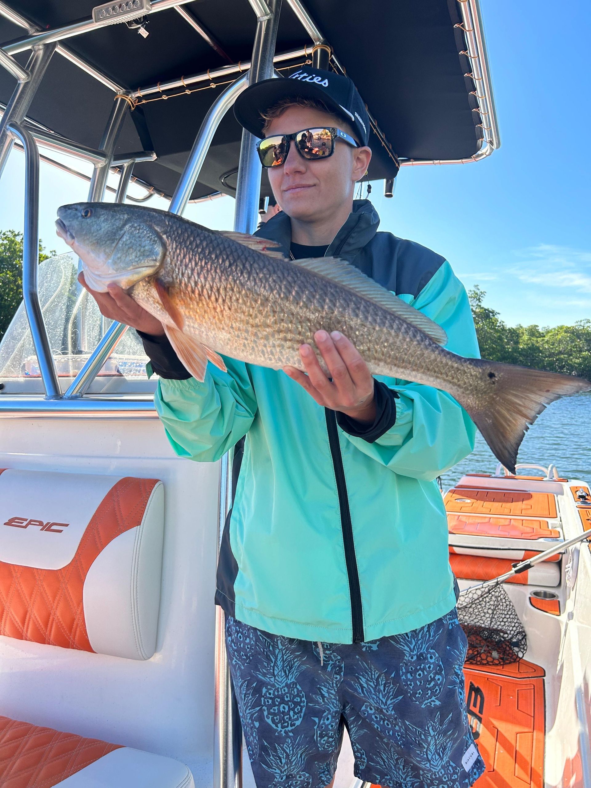 Person on a boat holds up a large redfish. They wear sunglasses, a jacket, and a hat. Bright sky and water.