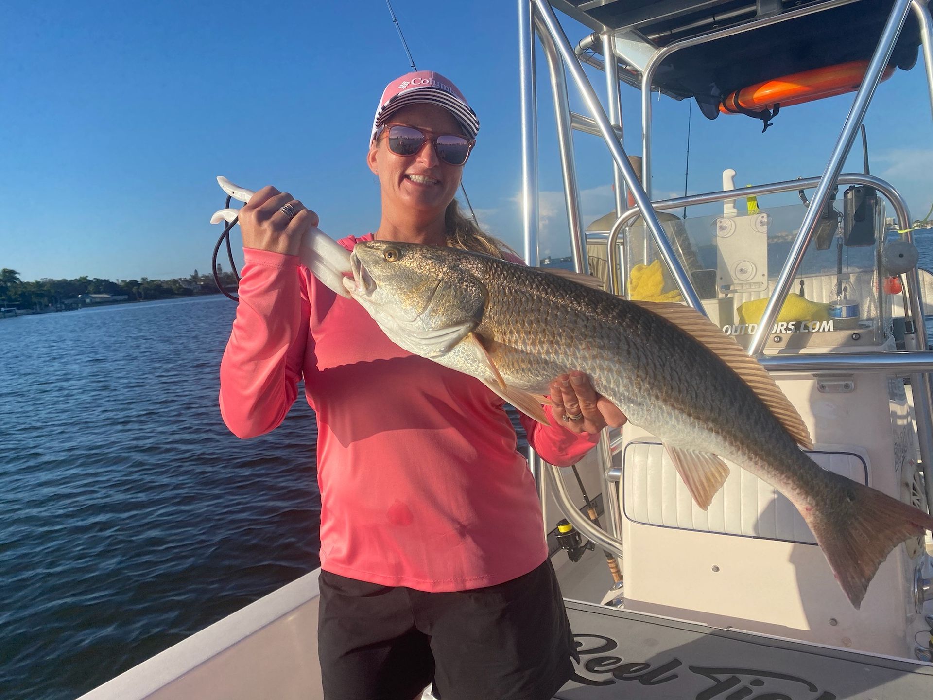 Woman on a boat holding up a large redfish. She wears pink shirt, sunglasses, and a hat. Blue water in background.