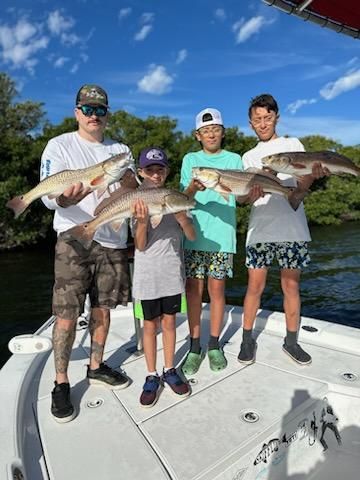 Group of four people on a boat holding fish. Blue sky, green trees, and the water are visible in the background.