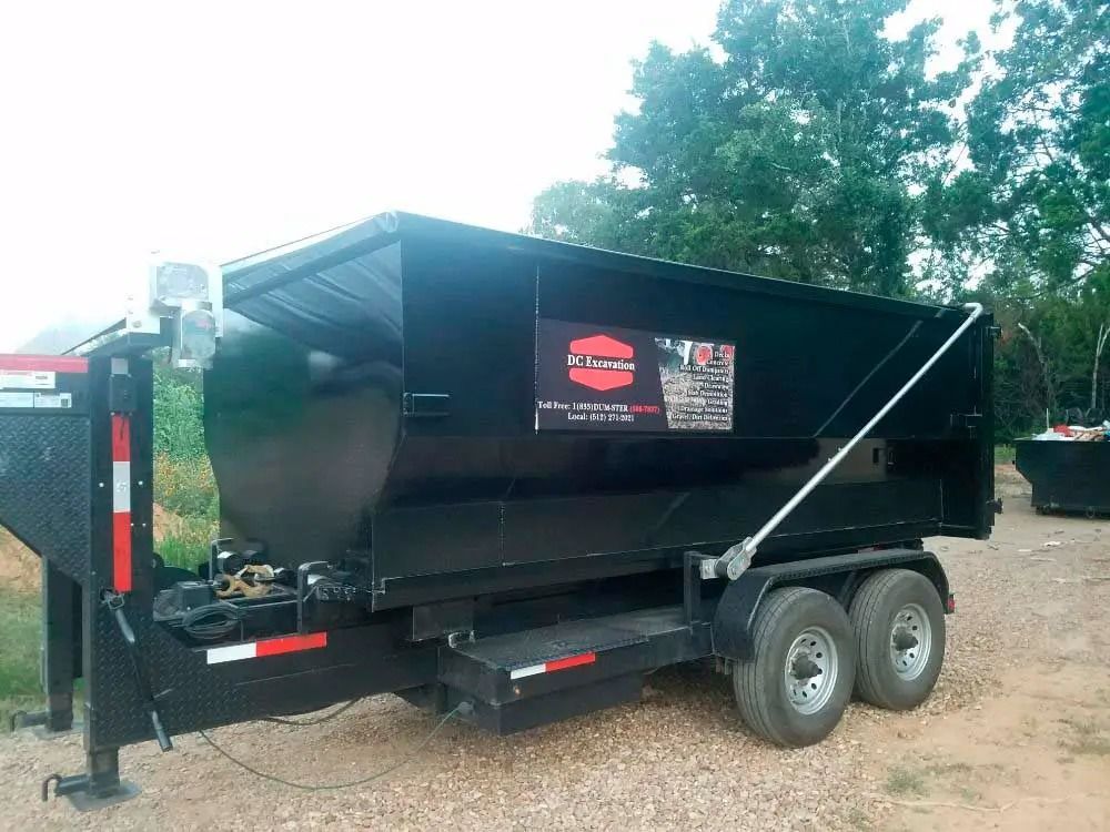 Black dump trailer with dual axles parked on dirt, showing company logos and a tarp cover.
