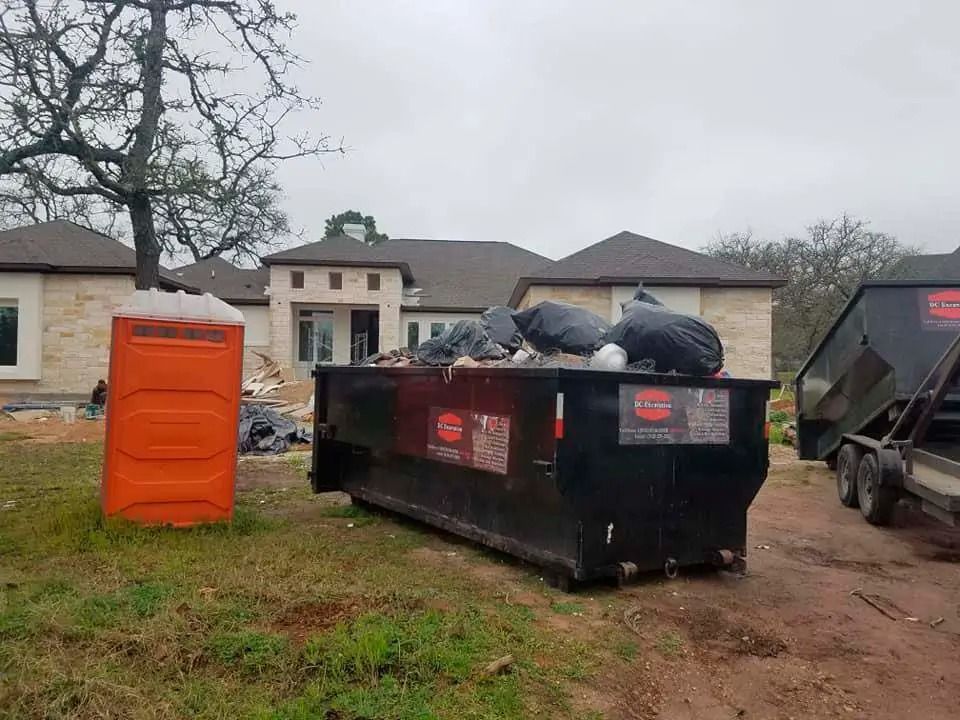 Orange portable toilet beside a black dumpster on a muddy lot with houses in the background