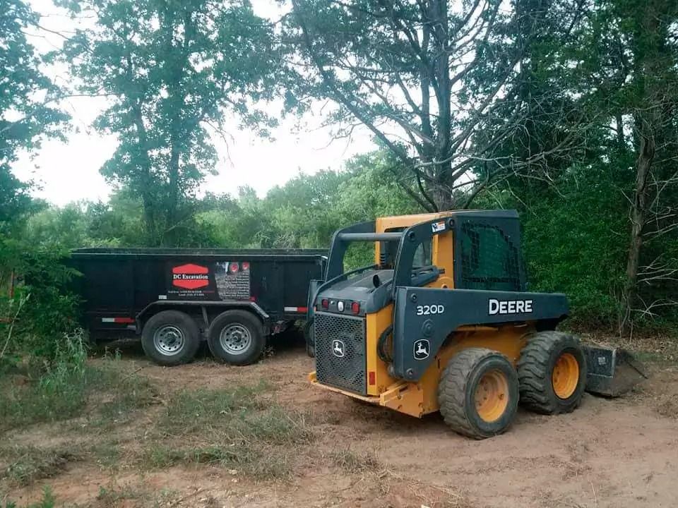 John Deere skid steer next to a black dump trailer in a grassy, wooded area